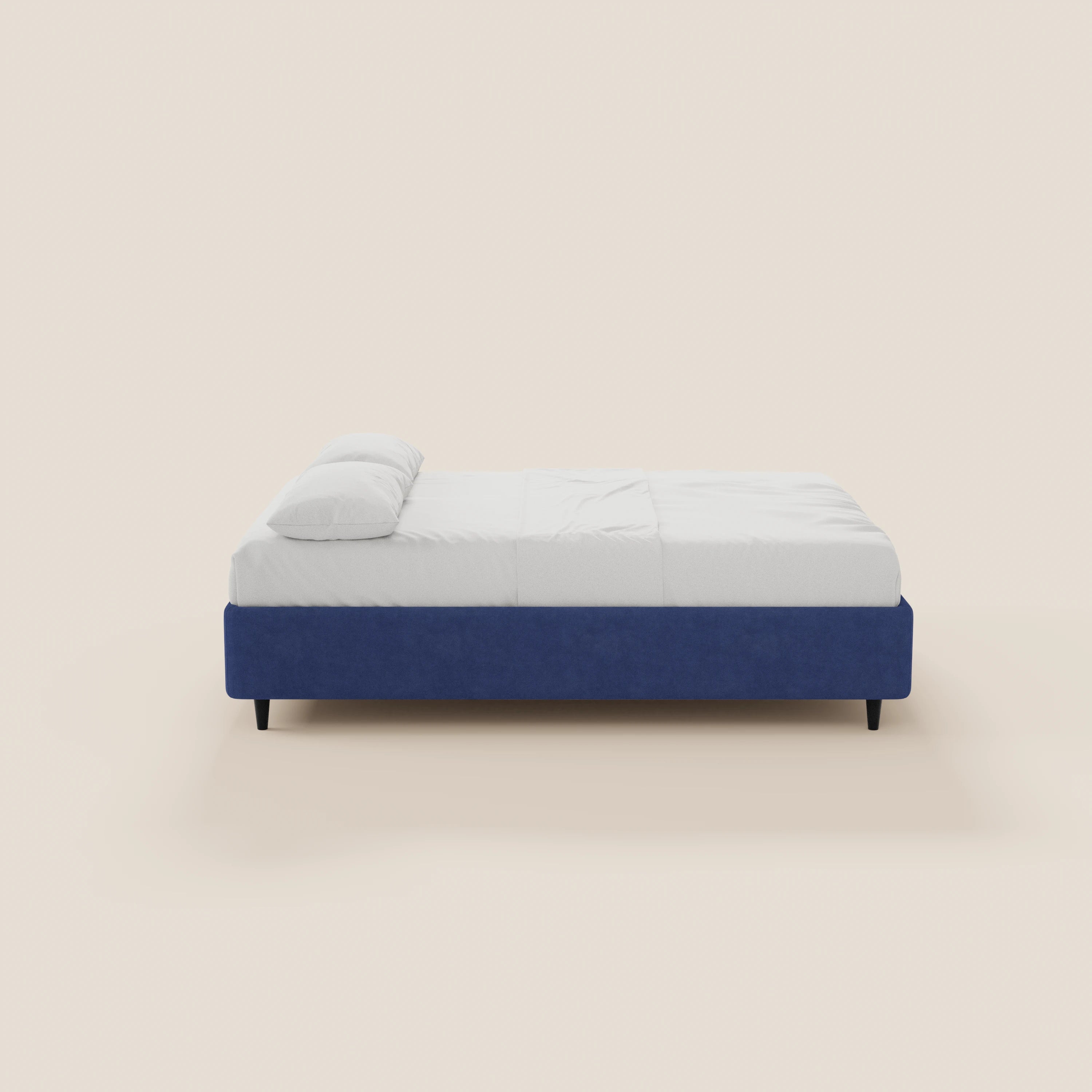 Una vista laterale del letto sommier ERON di Letti.store, caratterizzato da una base imbottita impermeabile Oeko-Tex di colore blu intenso con vano contenitore, biancheria da letto bianca, un cuscino singolo bianco, gambe corte scure e un design minimalista su uno sfondo beige chiaro.