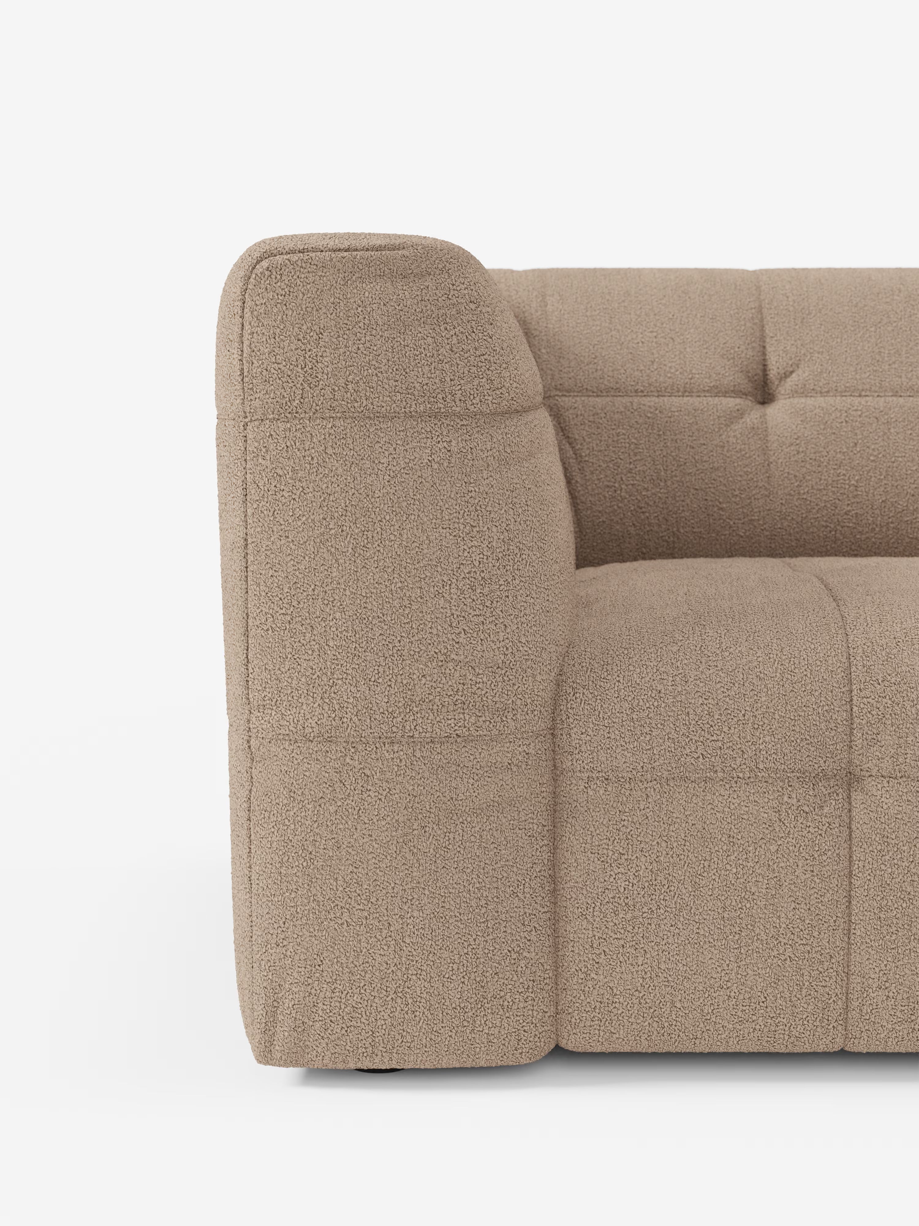 Un primo piano del bracciolo sinistro, della seduta e dello schienale del divano angolare componibile e modulare Mosaic in bouclé: capitonné e design in tessuto smacchiabile T21 di Divani.Store. Il rivestimento beige presenta un morbido tessuto bouclé con cuciture a vista e trapuntatura a quadrati, su uno sfondo bianco a tinta unita. Di questo elegante divano componibile è visibile solo una parte.