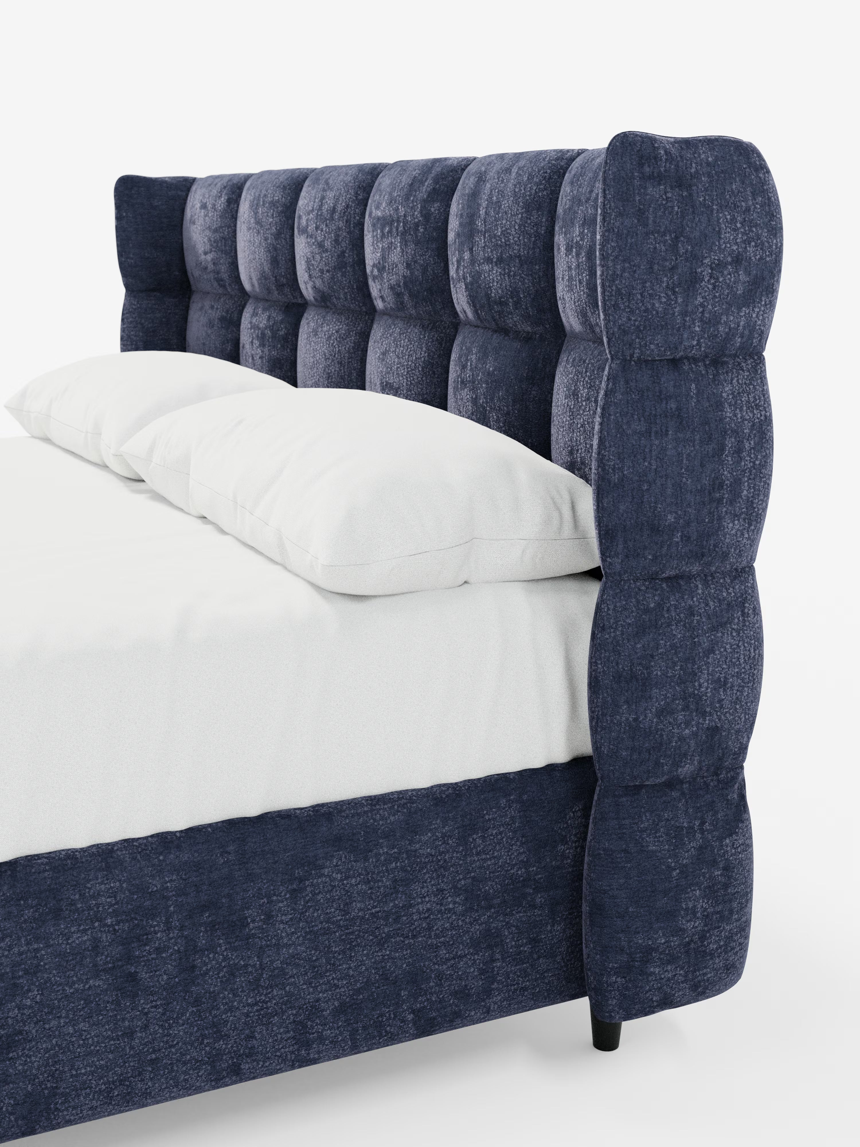 Primo piano del letto imbottito RAGNAR con contenitore e testiera capitonné avvolgente di Letti.store, caratterizzato da un’elegante testiera imbottita di colore blu intenso con finitura testurizzata e da una struttura in tessuto certificato Oeko-Tex®. La testiera capitonné presenta un motivo a griglia con morbide sezioni arrotondate. Sullo sfondo bianco a tinta unita si intravedono due cuscini bianchi e un lenzuolo bianco.