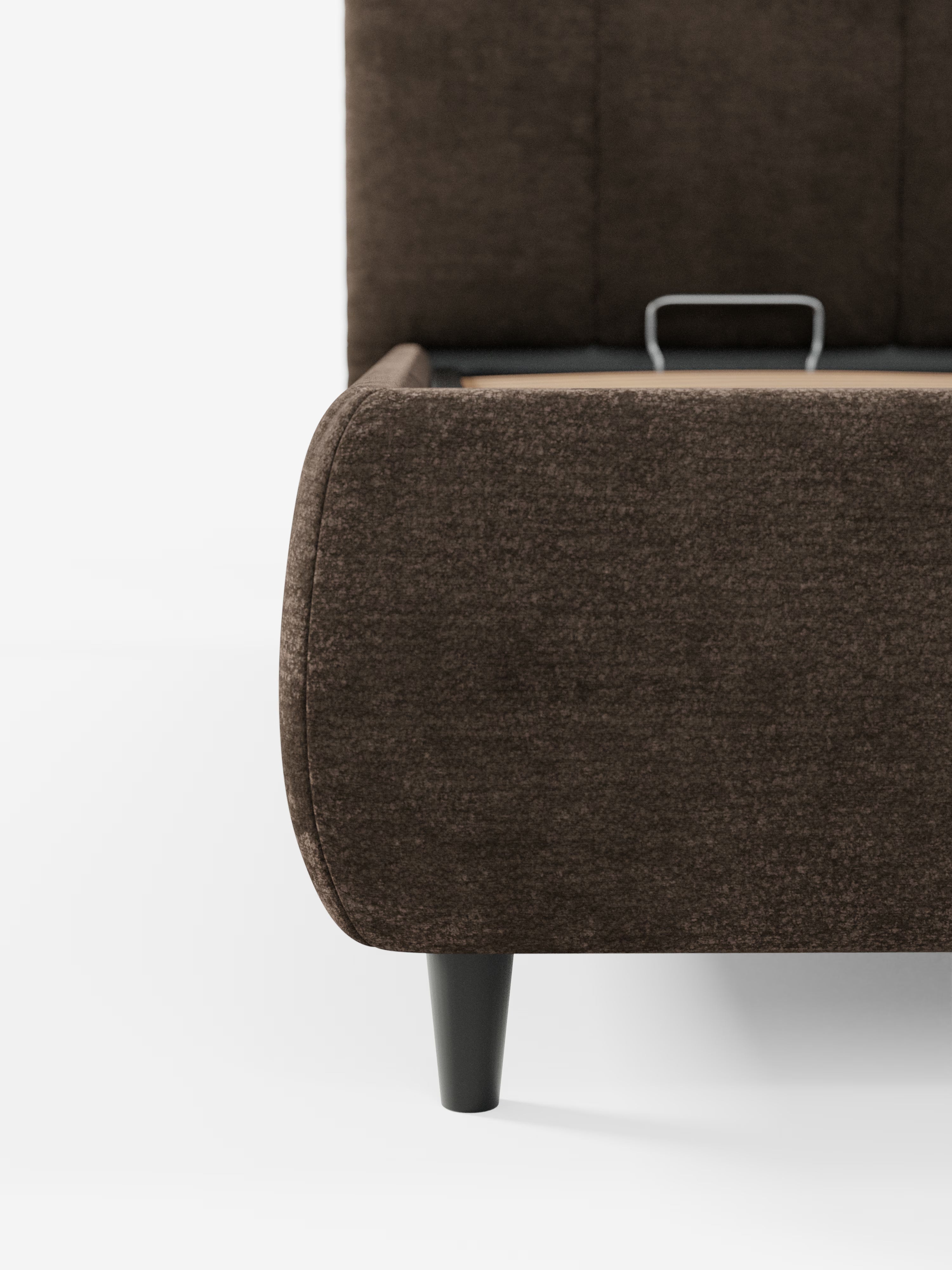 Un primo piano dell'angolo del letto imbottito ARKAM con contenitore e testiera a lavorazione verticale di Letti.store, caratterizzato da un rivestimento in tessuto marrone scuro certificato Oeko-Tex®, bordi arrotondati, una gamba affusolata nera e una maniglia metallica a vista sulla base del letto. Lo sfondo è bianco liscio.