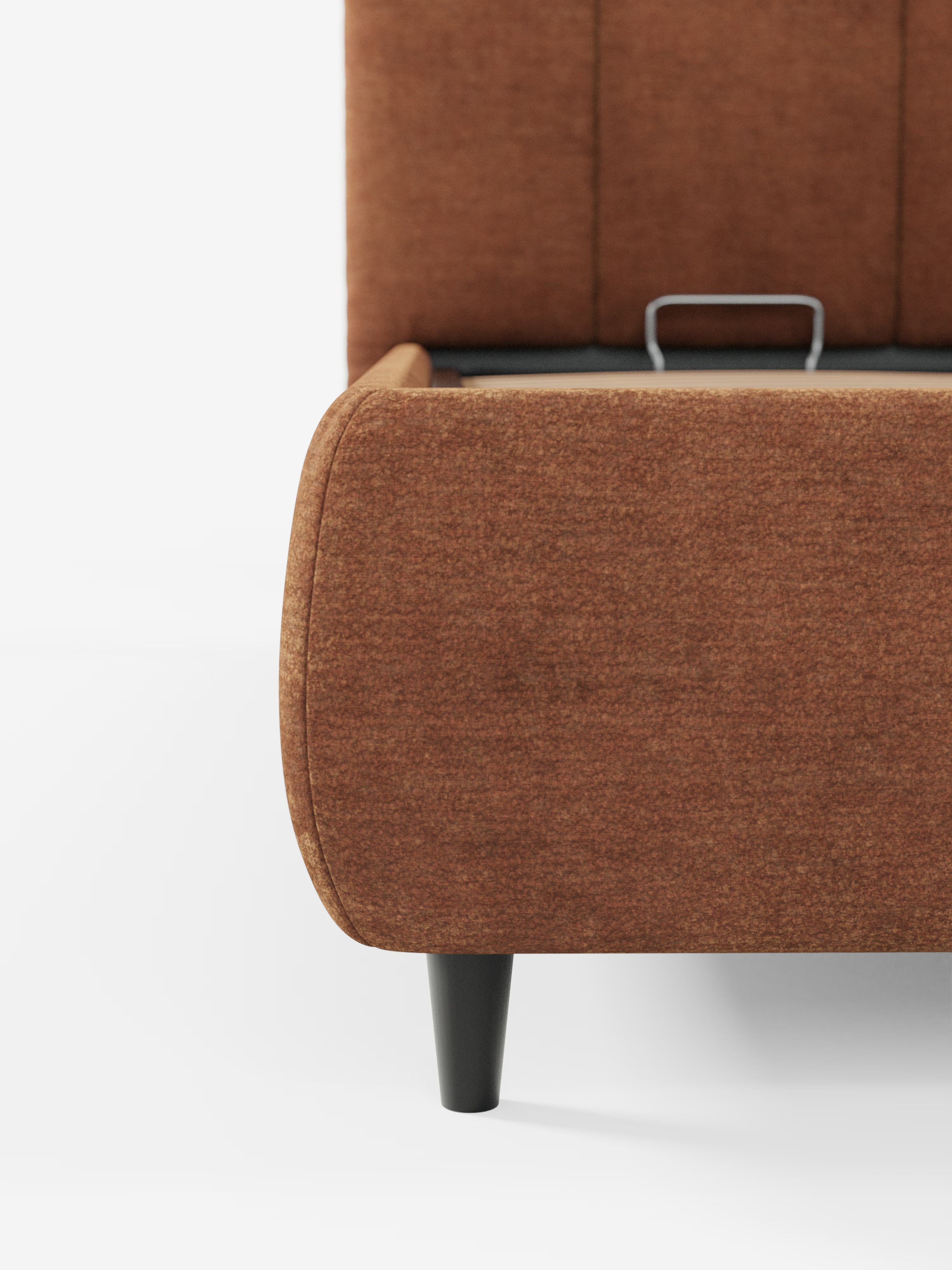 Primo piano dell'angolo in basso a destra del letto imbottito ARKAM con contenitore e testiera a lavorazione verticale di Letti.store, caratterizzato da un rivestimento in tessuto color ruggine certificato Oeko-Tex®, una gamba affusolata nera e una maniglia in metallo sulla rete a doghe. Parte della testiera imbottita con cuciture verticali è visibile su uno sfondo bianco a tinta unita.