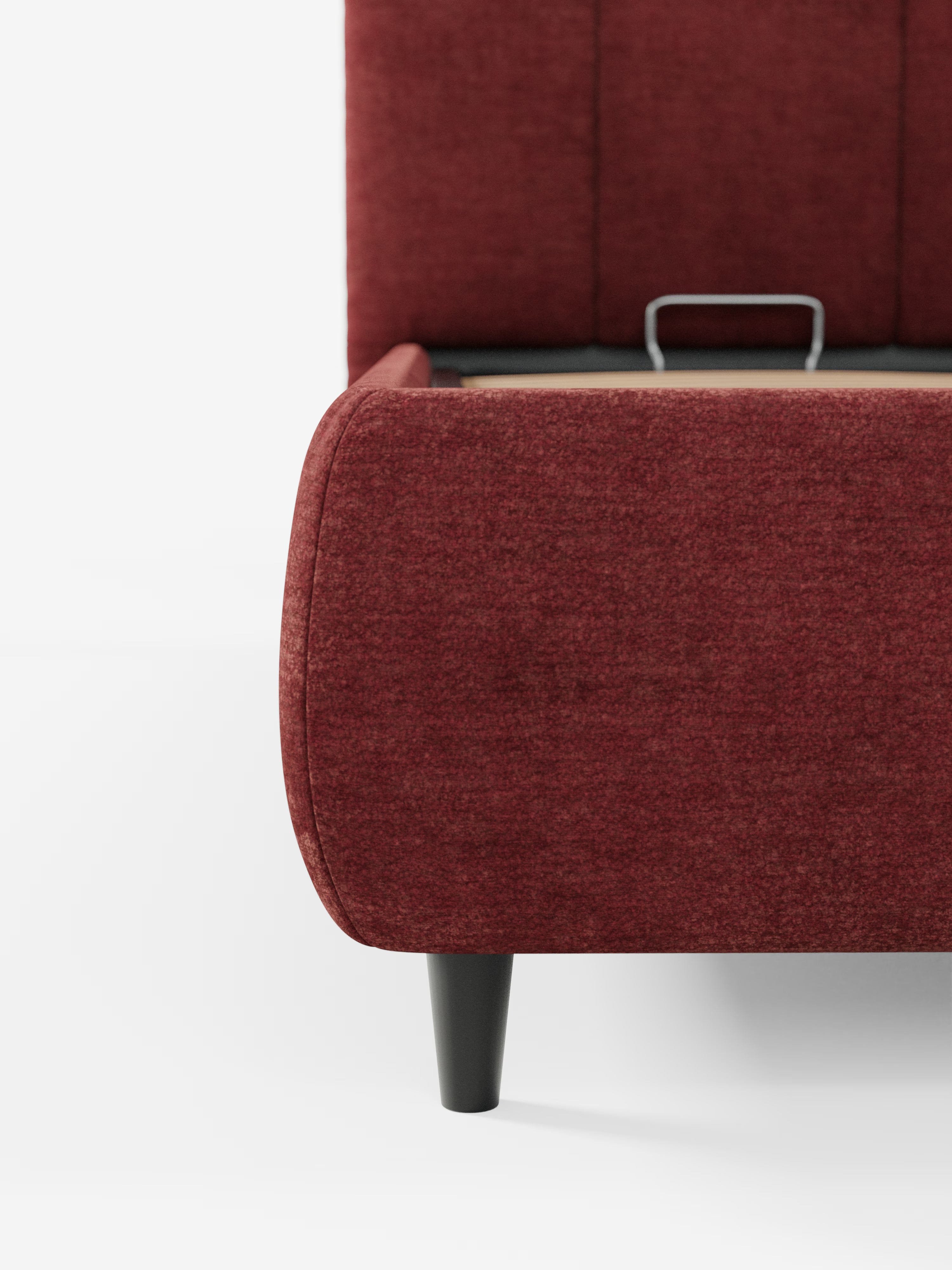 Primo piano dell'angolo inferiore del letto ARKAM imbottito con contenitore e testiera a lavorazione verticale di Letti.store, in rosso, con gambe nere. Il letto presenta un design morbido e sinuoso in stile shabby chic, realizzato in tessuto certificato Oeko-Tex®, e include un cassetto aperto — tipico di un letto contenitore — con una maniglia in metallo parzialmente visibile sul lato. Lo sfondo è bianco a tinta unita.