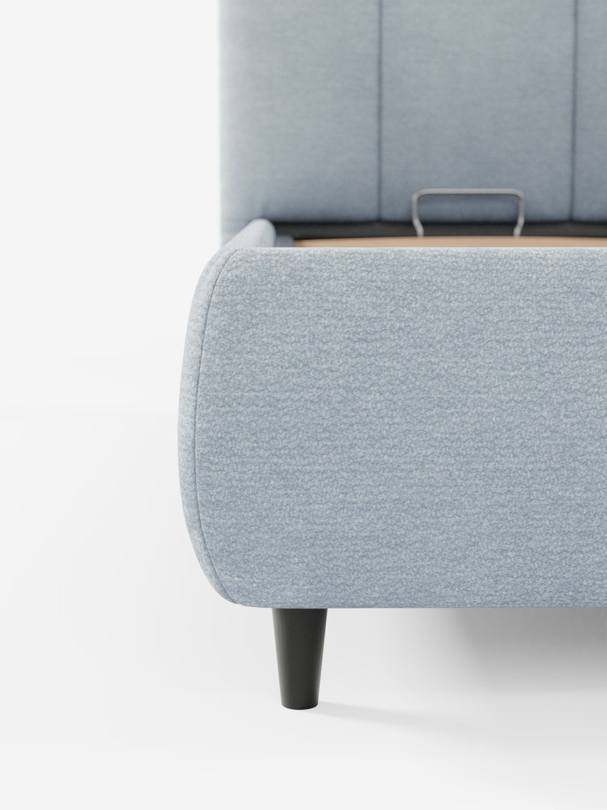 Primo piano del letto imbottito ARKAM con contenitore e testiera a lavorazione verticale di Letti.store, presentato in tessuto azzurro certificato Oeko-Tex®. L’immagine mette in risalto un angolo arrotondato, una gamba affusolata nera e una parte della testiera imbottita con cuciture verticali. Una maniglia in metallo alla base suggerisce la presenza di un comodo vano contenitore sotto il letto, caratteristica di questo pratico letto contenitore dal design shabby chic. Lo sfondo è bianco a tinta unita.