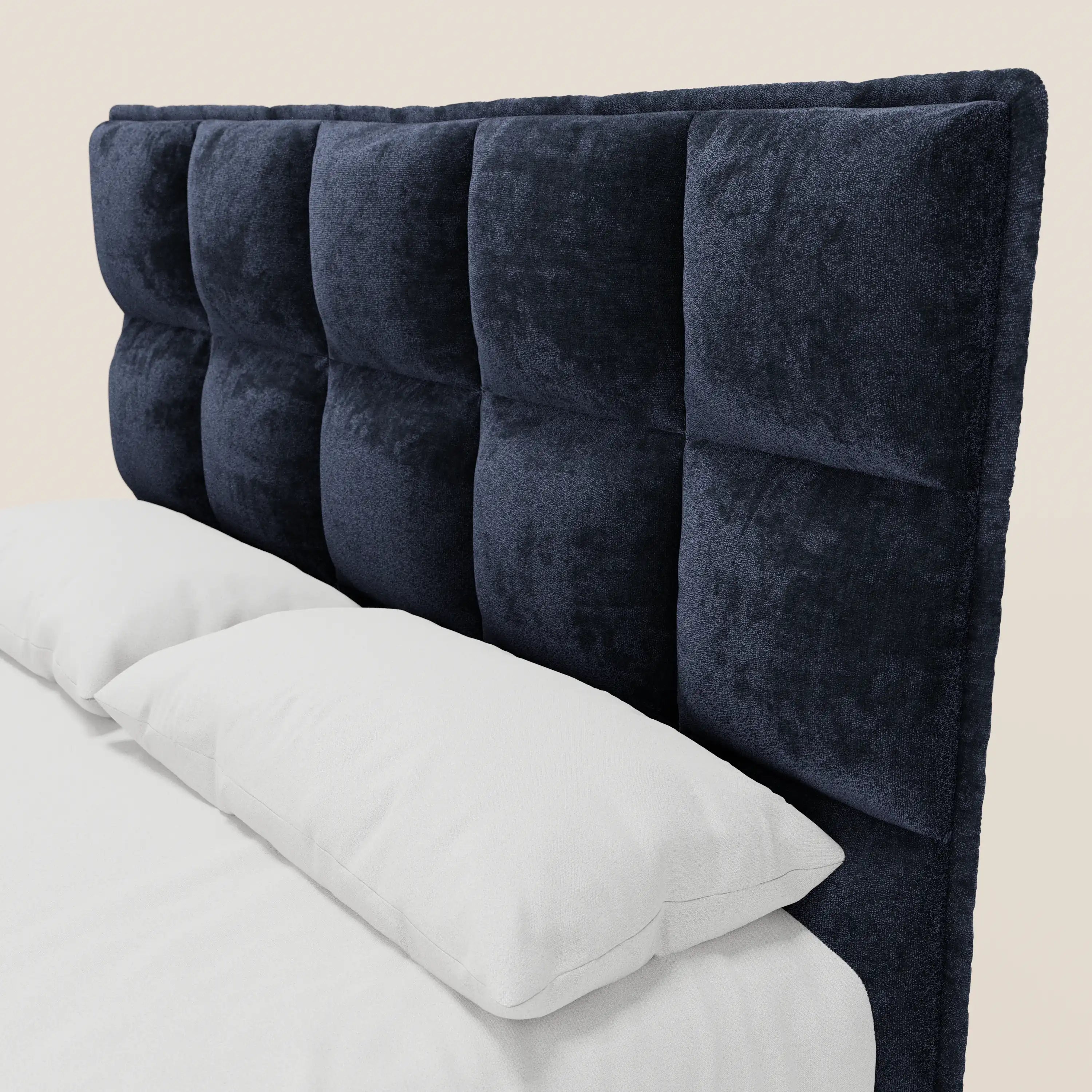 Un primo piano mostra il letto imbottito AURO di Letti.store, caratterizzato da un’alta e morbida testiera capitonné in ciniglia blu navy certificata Oeko-Tex®, con biancheria da letto e cuscini di un bianco candido, che risaltano sulla parete beige chiaro.