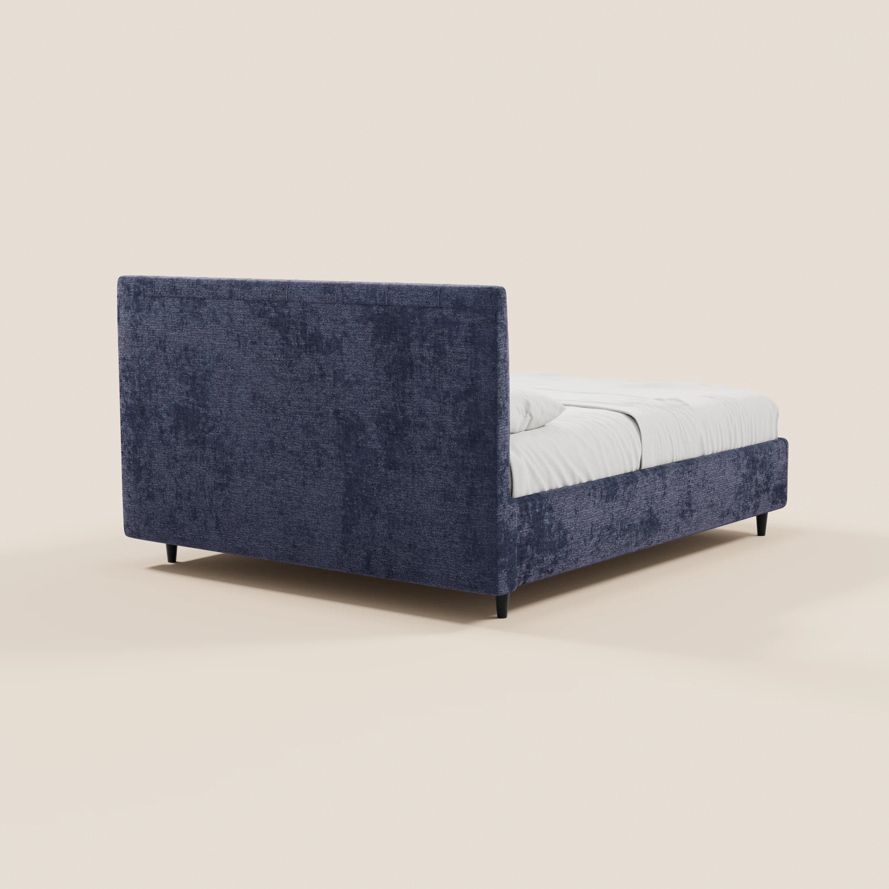 Il letto imbottito con contenitore ONYX di Letti.store presenta una struttura rivestita in morbido tessuto blu scuro certificato Oeko-Tex®, una testiera verticale imbottita, gambe basse nere e biancheria da letto bianca. L'immagine mostra il letto visto da un angolo posteriore su uno sfondo beige chiaro a tinta unita. Non sono visibili altri oggetti.