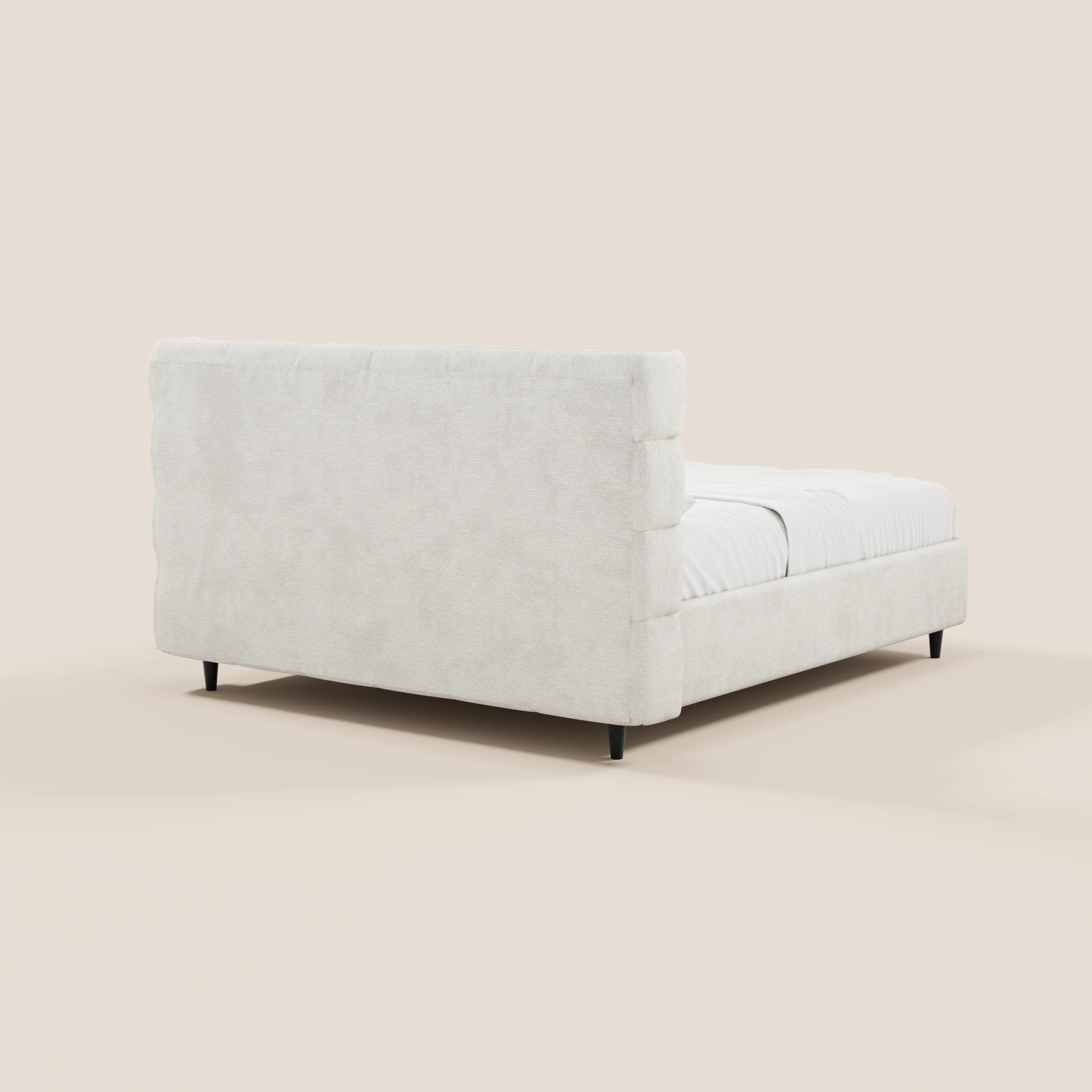 Il letto imbottito RAGNAR di Letti.store, con vano contenitore e testiera capitonné avvolgente in elegante tessuto Oeko-Tex®, è raffigurato nella vista dall’angolo posteriore sinistro, con rivestimento color crema, lenzuolo bianco e gambe nere su sfondo beige.