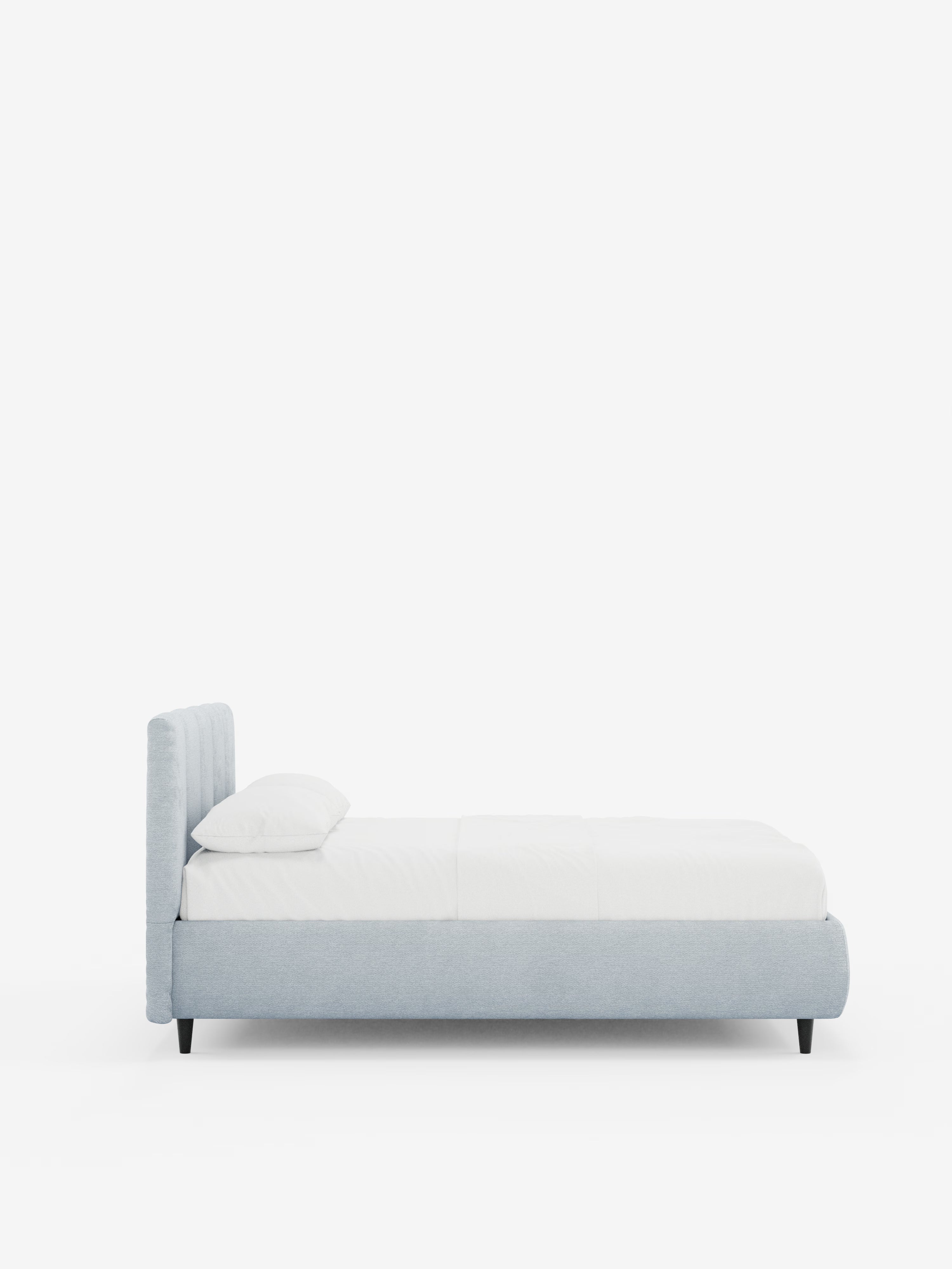 Vista laterale del letto imbottito ARKAM con contenitore e testiera a lavorazione verticale di Letti.store, caratterizzato da struttura e testiera in tessuto grigio chiaro certificato Oeko-Tex®, biancheria da letto bianco candido con un unico cuscino e quattro gambe corte nere. Lo sfondo bianco a tinta unita mette in risalto il design shabby chic moderno di questo elegante letto imbottito.