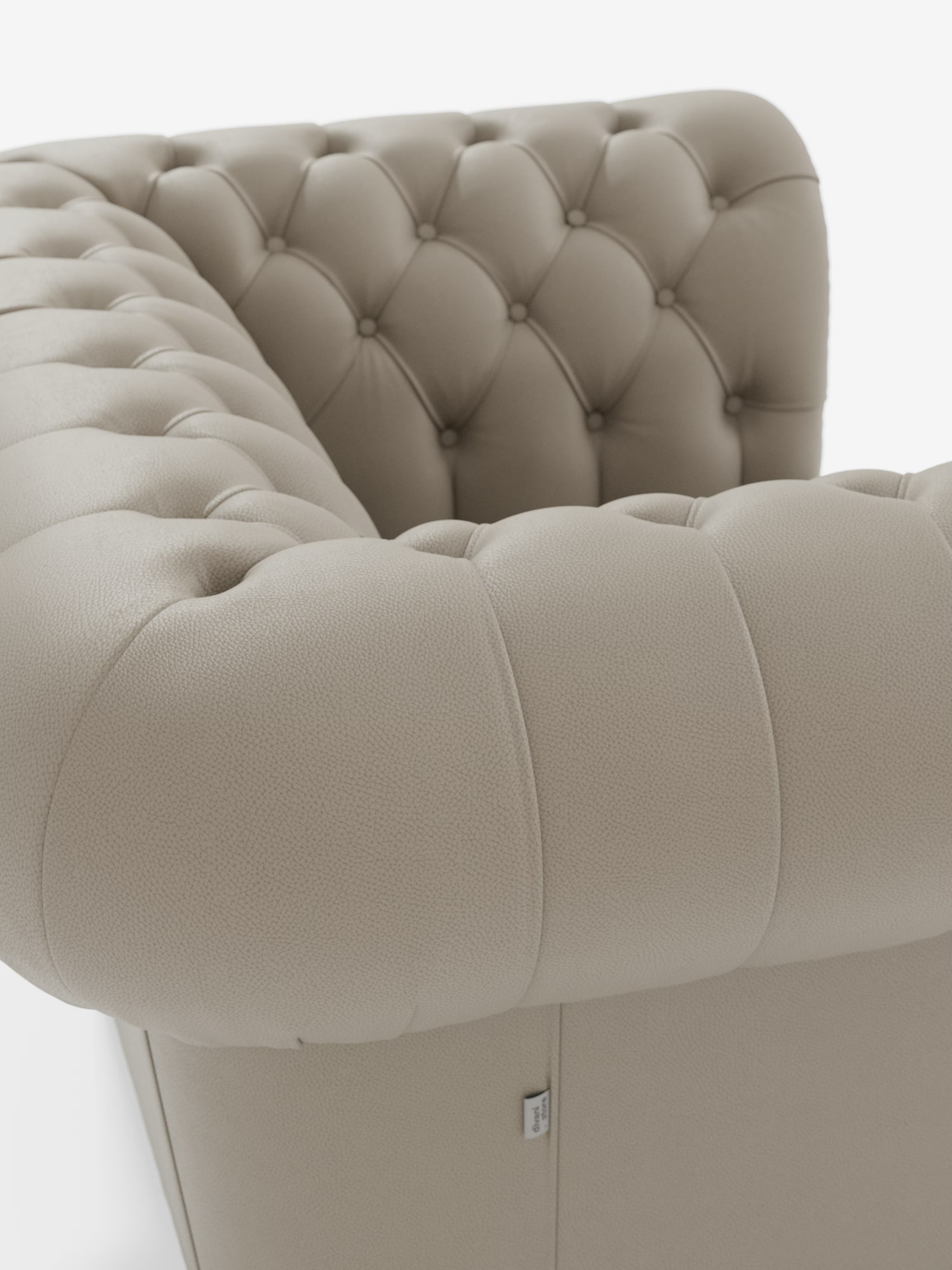 Primo piano dell'angolo della poltrona Chester in vera pelle spessorata con capitonné artigianale di Divani.Store, che mette in risalto il rivestimento in vera pelle beige con una profonda trapuntatura artigianale a bottoni. Il design classico — che richiama gli iconici divani Chester — presenta braccioli spessi e arrotondati e un'etichetta in tessuto visibile vicino alla base. La meticolosa lavorazione artigianale è evidente in ogni punto, il tutto su uno sfondo bianco e pulito da studio.