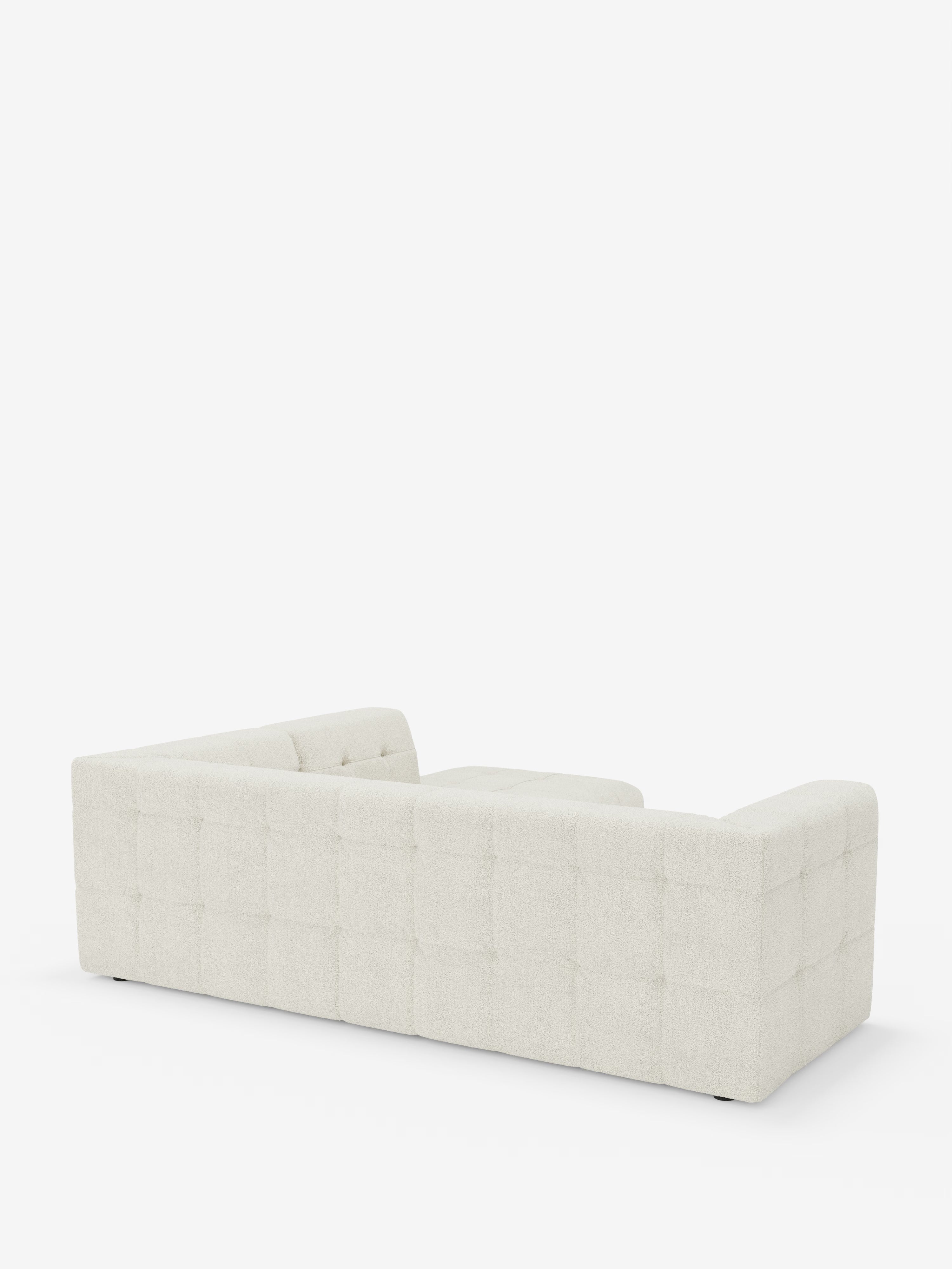 Un divano bianco moderno e dal profilo basso visto da dietro: il divano angolare componibile e modulare Mosaic in bouclé di Divani.Store presenta una silhouette squadrata, una seduta profonda e un rivestimento capitonné trapuntato con motivo a griglia. Perfetto per un soggiorno moderno, questo versatile divano componibile è presentato su uno sfondo bianco a tinta unita per mettere in risalto le sue linee pulite e il design contemporaneo in tessuto smacchiabile T21.