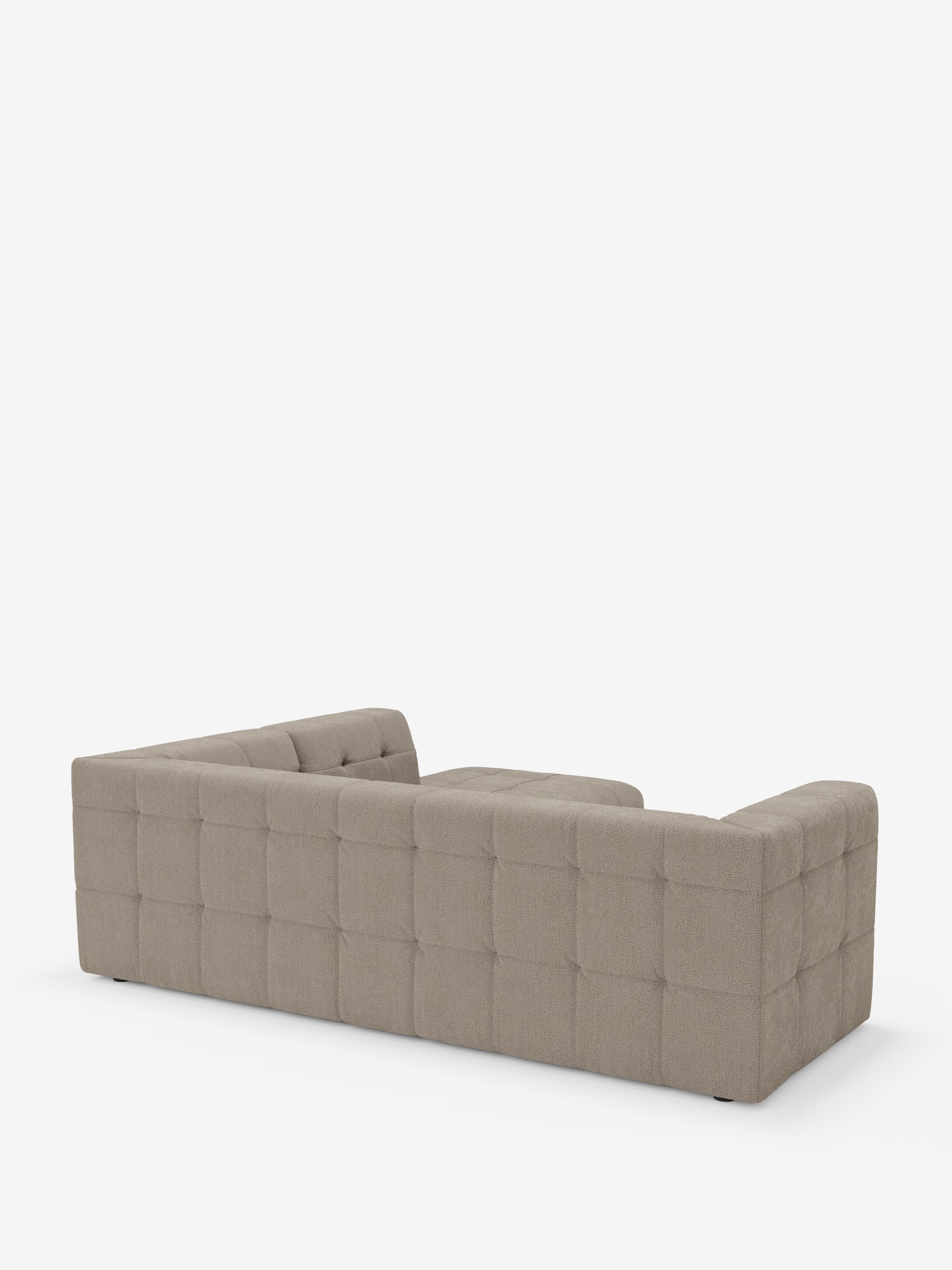 Un divano angolare componibile e modulare Mosaic, moderno e dal profilo ribassato, in bouclé beige di Divani.Store, caratterizzato da una profonda trapuntatura capitonné sullo schienale e sui fianchi, è ripreso di profilo da dietro su uno sfondo bianco. Questo divano modulare presenta un design squadrato e simmetrico senza gambe a vista ed è rivestito in tessuto T21 resistente alle macchie.