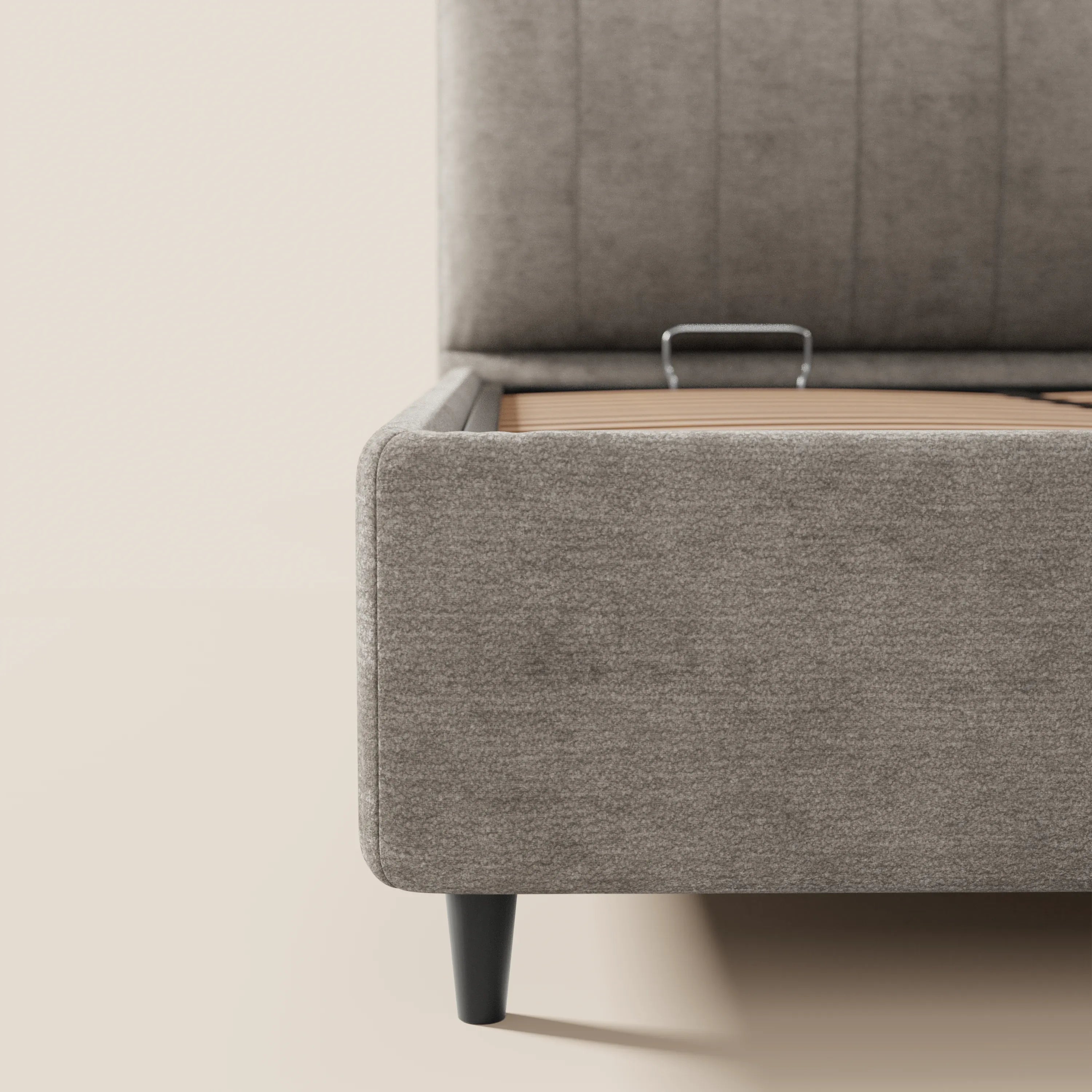 Un primo piano del letto imbottito ONYX con contenitore di Letti.store, realizzato in tessuto grigio chiaro certificato Oeko-Tex®, che mette in risalto le gambe affusolate nere e la base rialzata che rivela lo spazio contenitivo, su sfondo di pareti e pavimento beige neutro.