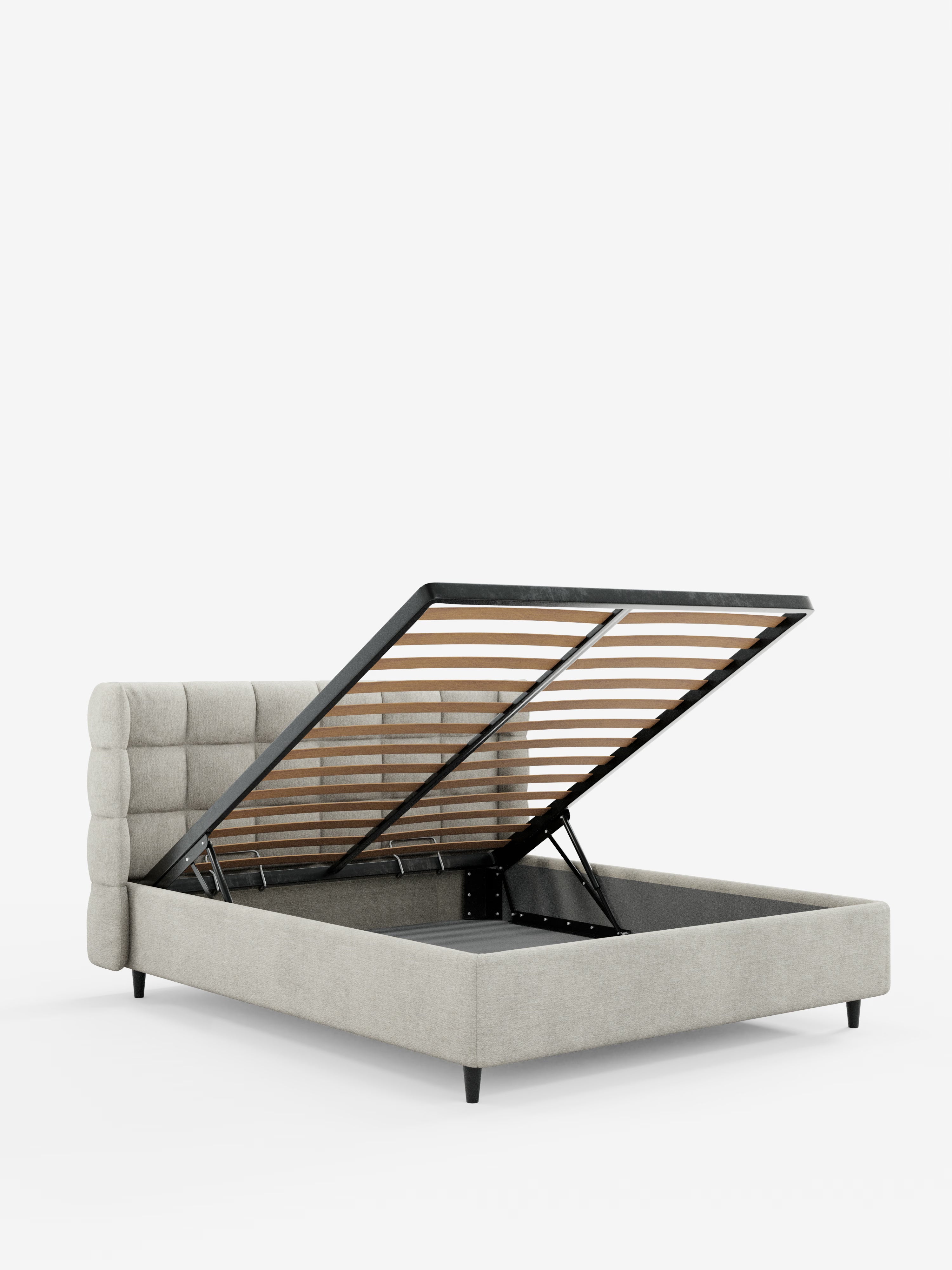Un letto imbottito RAGNAR grigio chiaro con vano contenitore e testiera capitonné avvolgente di Letti.store, realizzato con un elegante tessuto certificato Oeko-Tex® e dotato di gambe nere. La base a doghe del materasso si solleva per rivelare un ampio vano contenitore nascosto al di sotto. Fotografato su uno sfondo bianco a tinta unita.