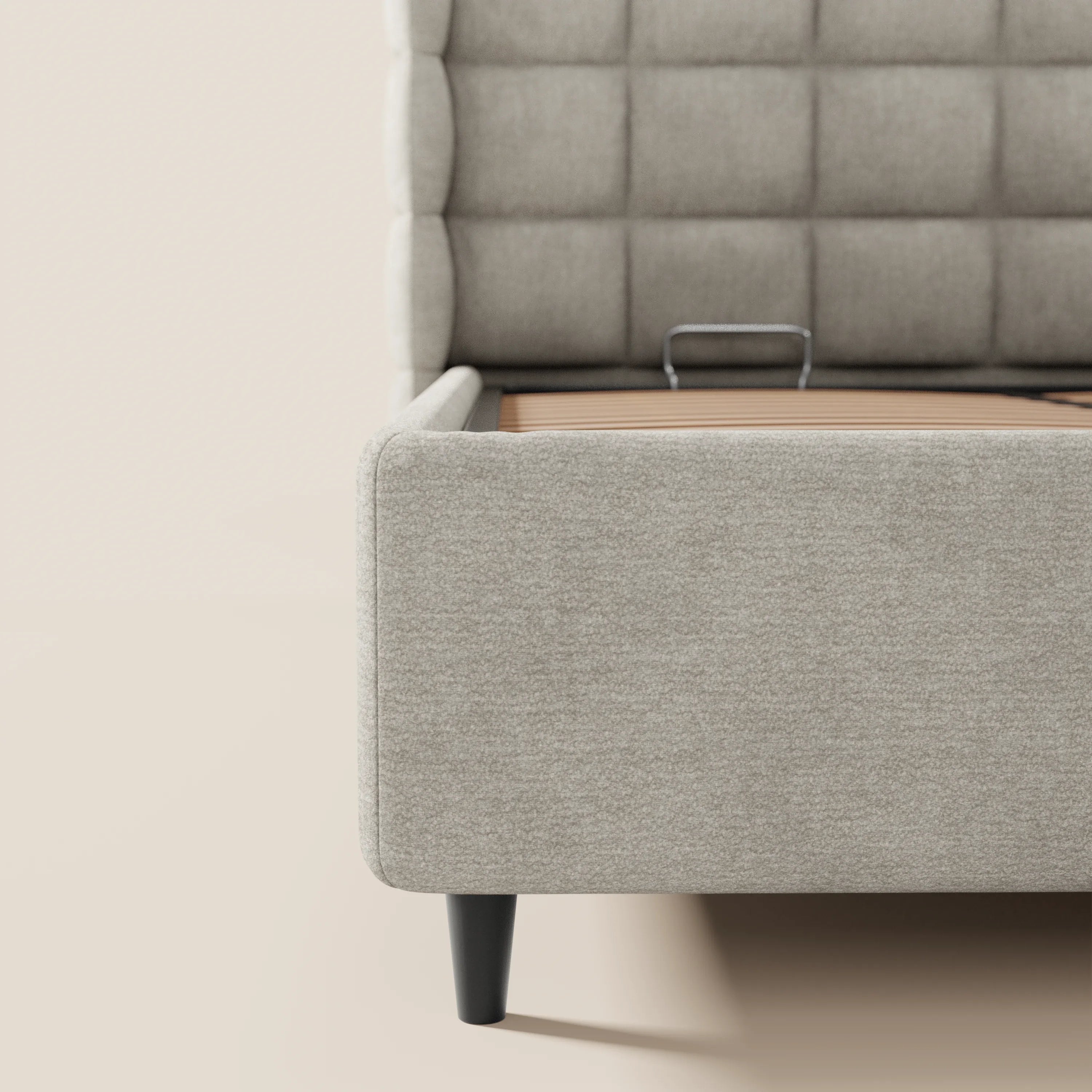 Primo piano del lato destro del letto imbottito RAGNAR di Letti.store, in tessuto beige chiaro certificato Oeko-Tex®, con testiera avvolgente capitonné, gambe affusolate nere e un accenno al meccanismo del contenitore. Sfondo beige chiaro.