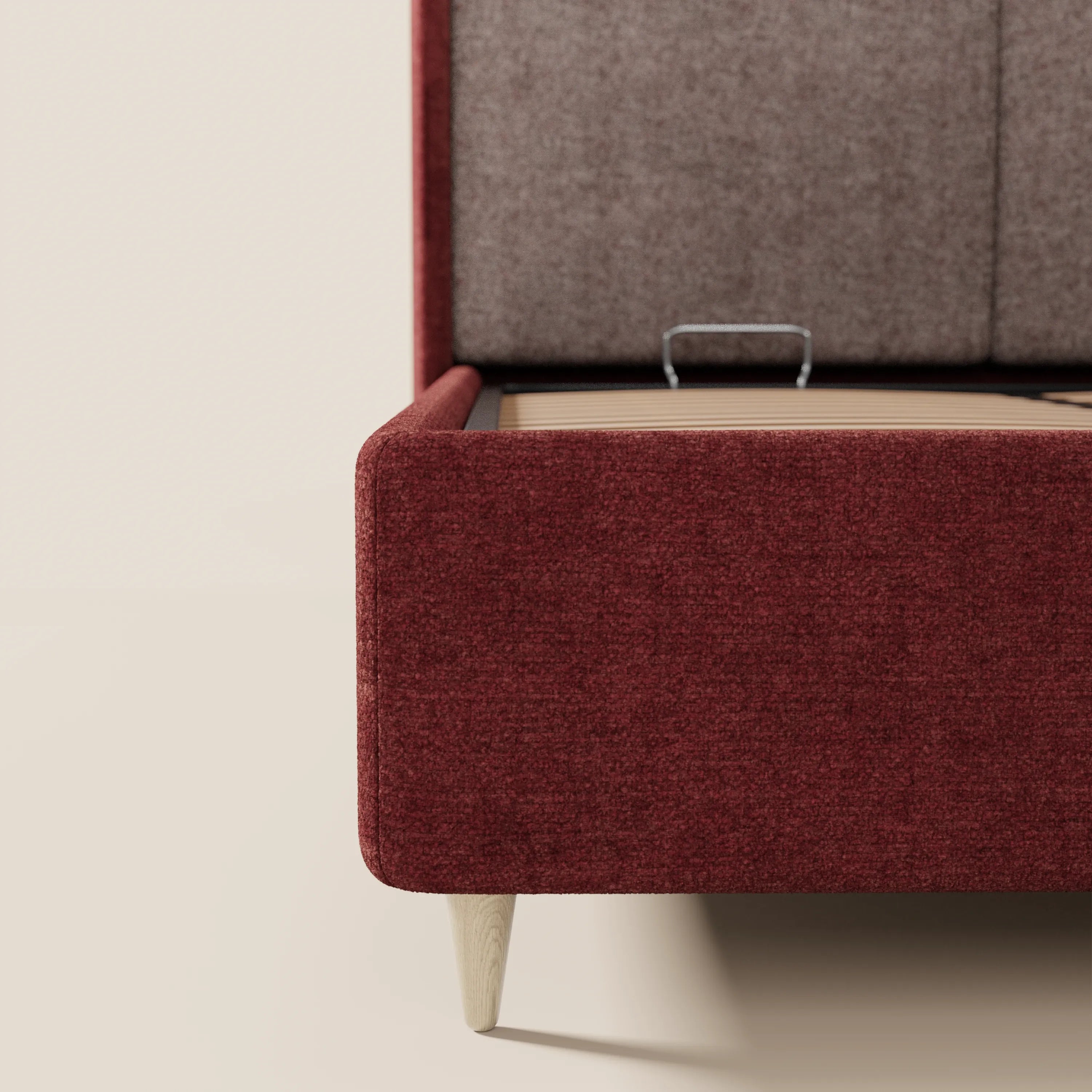 Primo piano del letto imbottito bicolore VALKIRIA di Letti.store, caratterizzato da un tessuto rosso intenso, gambe in legno chiaro e un vano portaoggetti aperto con interno in legno e maniglia in metallo. Arredamento minimalista con sfondo e pavimento beige.