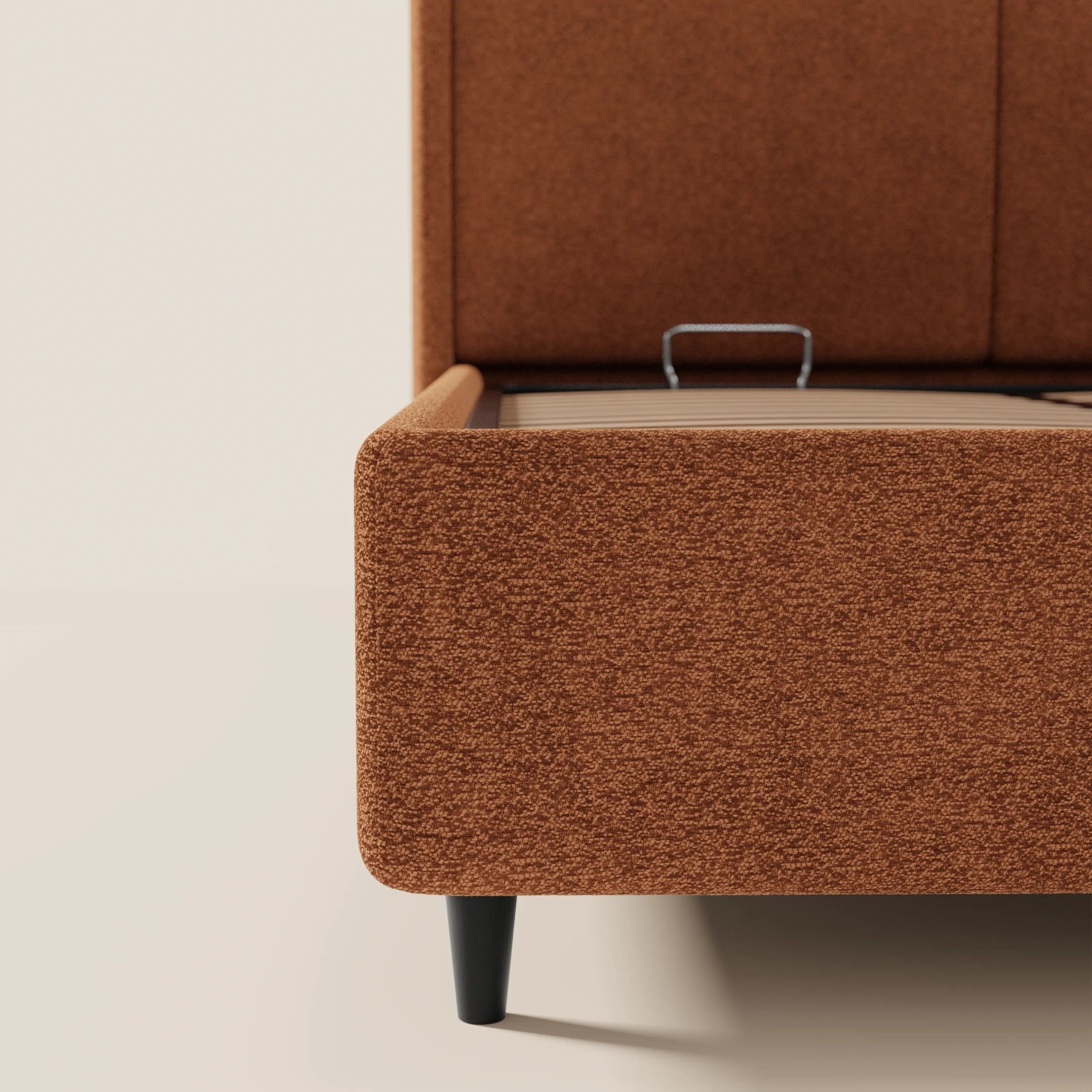 Primo piano del letto di design VALKIRIA di Letti.store, realizzato in tessuto ruggine testurizzato certificato Oeko-Tex®, dotato di 2 cuscini imbottiti, gambe cilindriche nere e un vano portaoggetti. Il design contemporaneo della base e dei fianchi risalta sullo sfondo di una parete dai toni neutri.