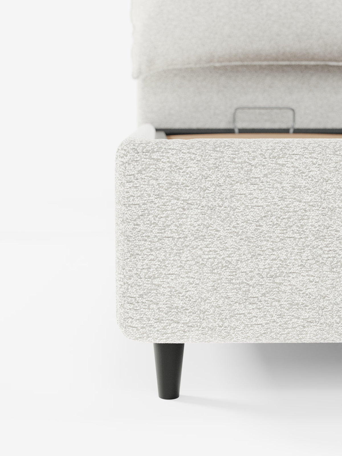 Primo piano di un letto imbottito ZAYRON, dalla trama ruvida e di colore grigio chiaro, con vano contenitore e morbidi cuscini regolabili, realizzato da Letti.store, caratterizzato da un moderno tessuto certificato Oeko-Tex®. L'angolo mostra gambe affusolate nere e una staffa di sostegno in metallo a vista all'interno del telaio. Il tutto su uno sfondo bianco a tinta unita che mette in risalto il tessuto del letto e i dettagli costruttivi. È visibile solo una parte del letto.