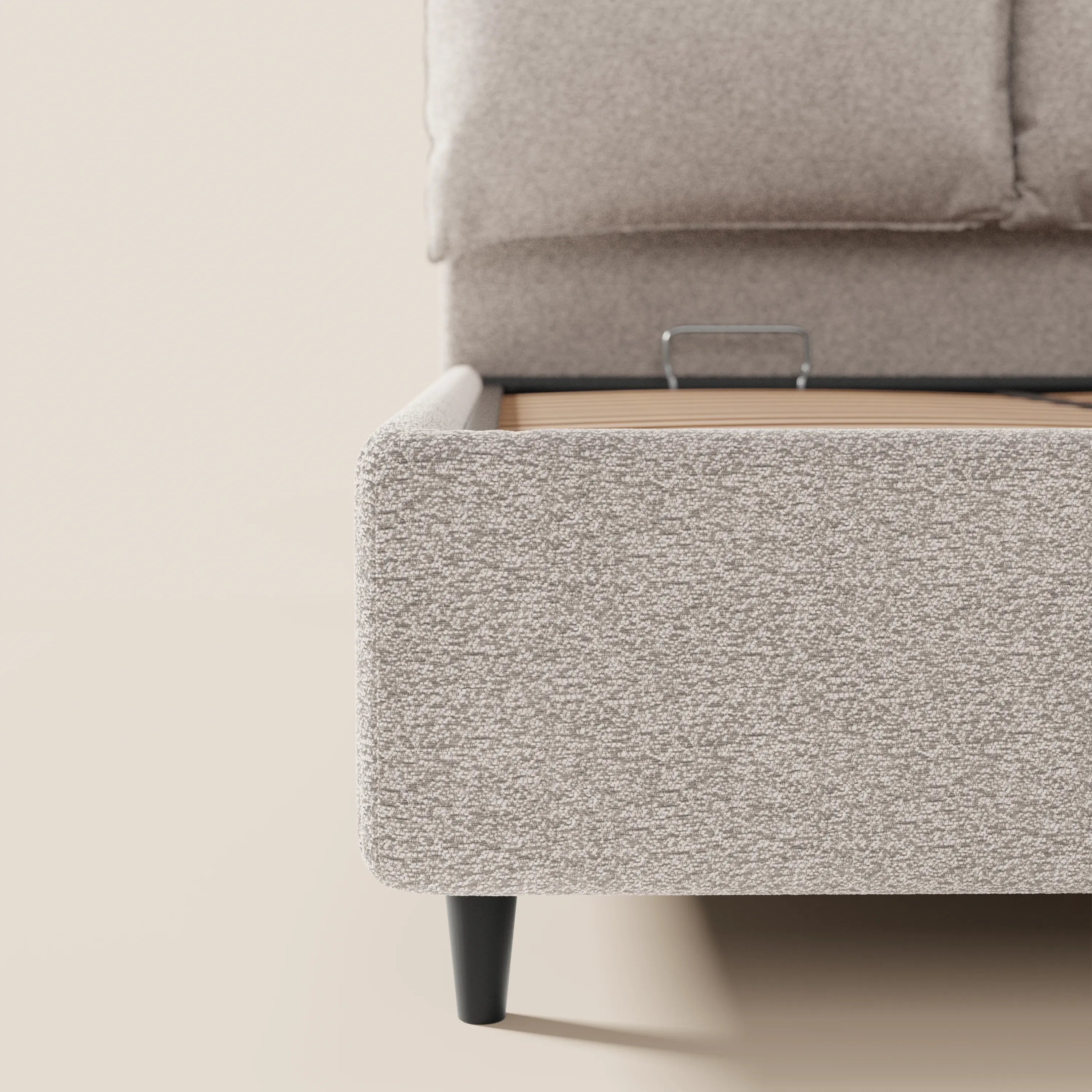 Primo piano del letto imbottito ZAYRON di Letti.store: tessuto beige chiaro certificato Oeko-Tex®, gambe angolari in legno nero, testiera imbottita regolabile, doghe e vano contenitore nascosto. Sfondo e pavimento in tonalità neutre chiare abbinate.
