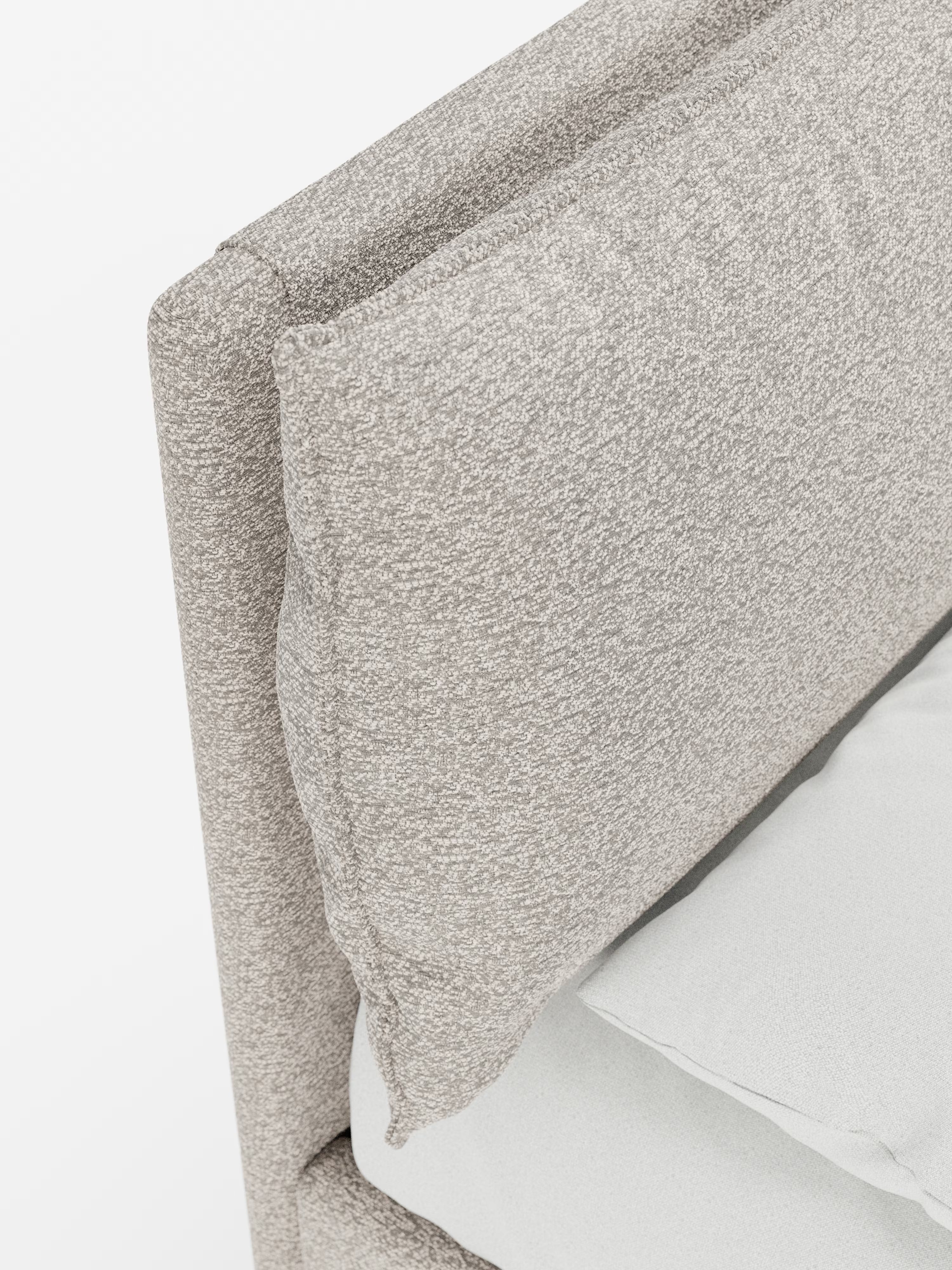 Primo piano del letto imbottito ZAYRON con contenitore e morbidi cuscini regolabili di Letti.store, che presenta una testiera contemporanea in tessuto certificato Oeko-Tex® in una tonalità di beige chiaro, morbida e dalla trama particolare, con un cuscino spesso appoggiato contro di essa. La biancheria da letto è di colore grigio chiaro, il tutto su uno sfondo bianco a tinta unita.
