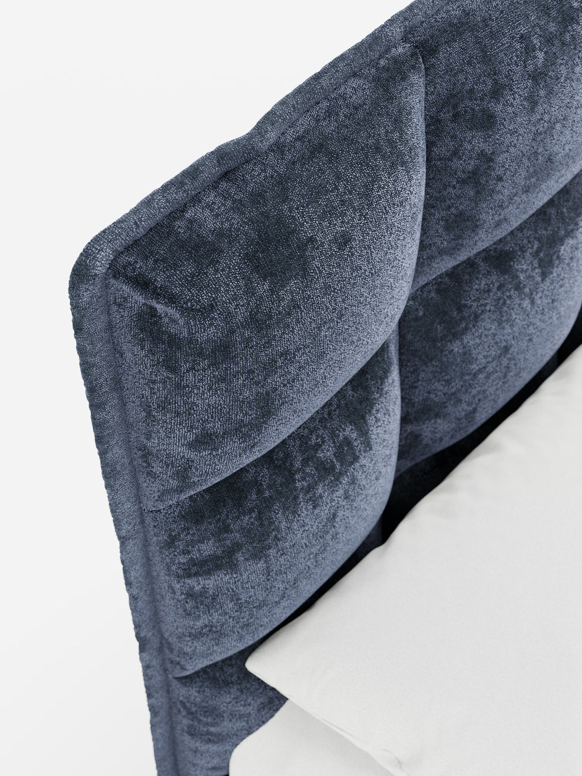 Primo piano dell'angolo del letto imbottito AURO di Letti.store, caratterizzato da un morbido rivestimento in ciniglia blu scuro certificato Oeko-Tex® e da una testiera alta, capitonné, con imbottitura a quadrati. Un cuscino bianco è appoggiato alla parte inferiore di questo elegante letto moderno con contenitore. Lo sfondo è semplice e ben illuminato.