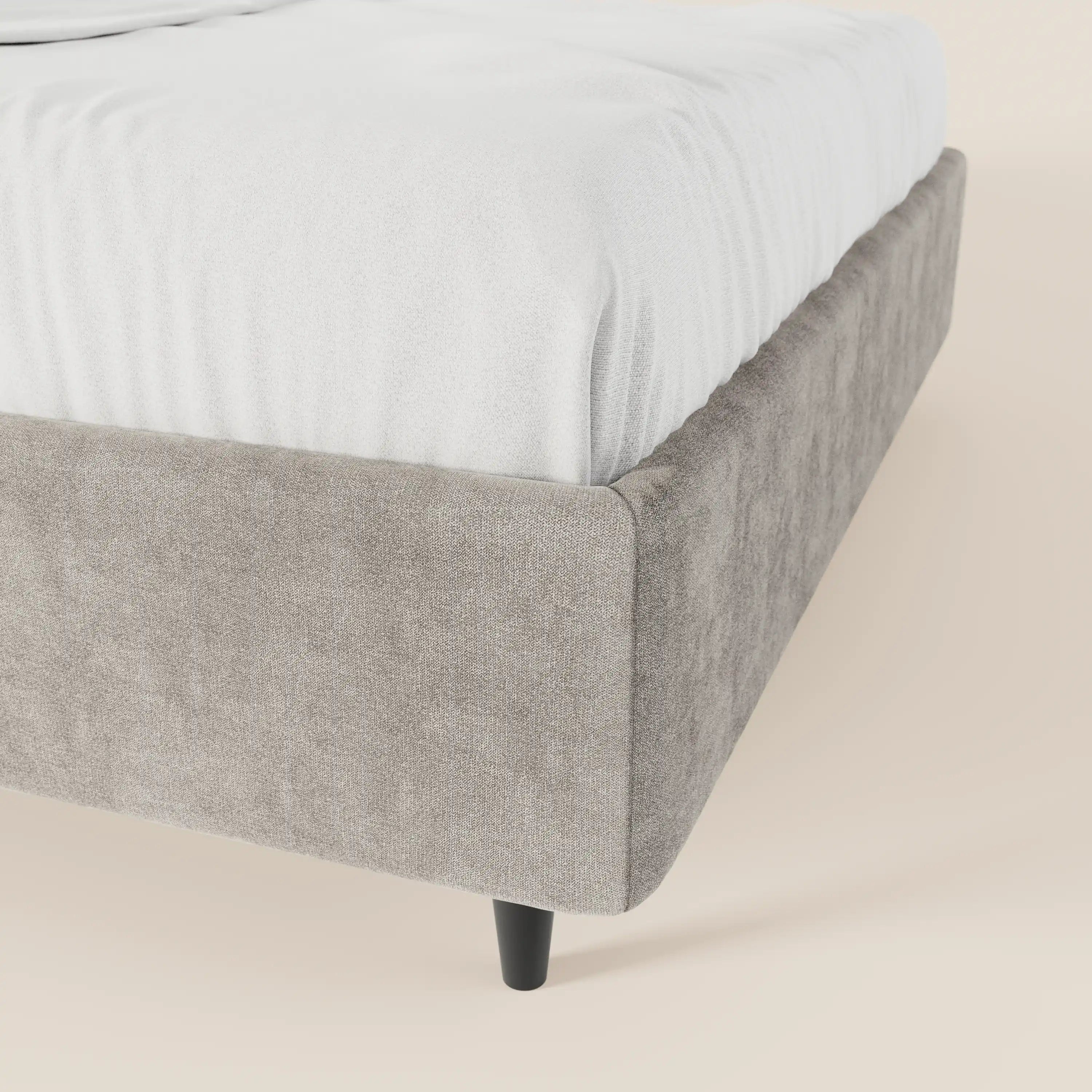 Un primo piano del letto sommier ERON di Letti.store mette in risalto il suo tessuto impermeabile grigio chiaro certificato Oeko-Tex, la morbida consistenza e le gambe affusolate nere che sostengono un materasso bianco. Lo sfondo presenta una superficie liscia di colore beige chiaro.