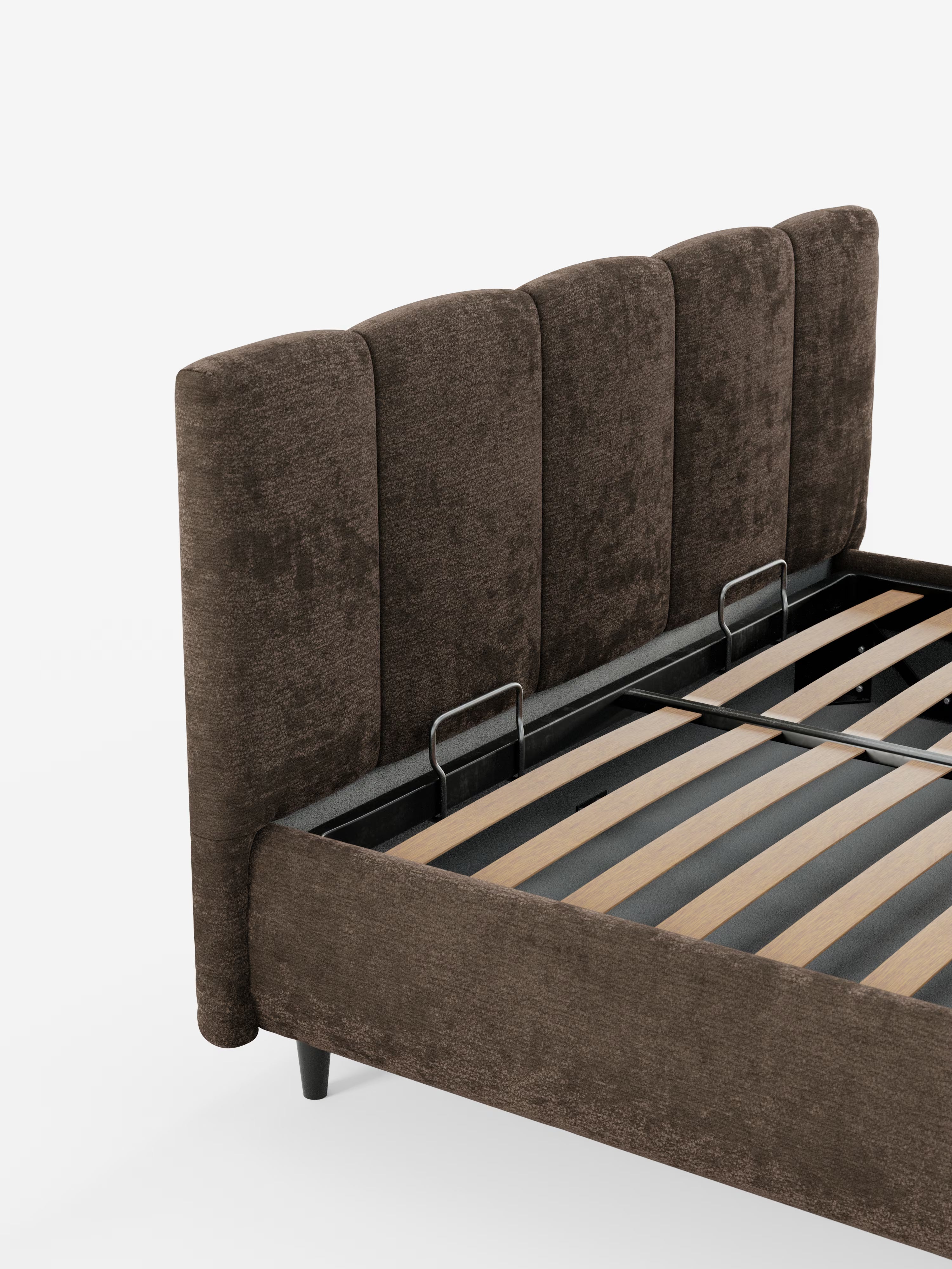 Un primo piano del letto imbottito ARKAM con contenitore e testiera a cuciture verticali di Letti.store mette in risalto una struttura del letto rivestita in marrone con una testiera alta, caratterizzata da cuciture verticali, realizzata in tessuto certificato Oeko-Tex®. Questo letto moderno in stile shabby chic presenta doghe in legno a vista, due barre di sostegno in metallo e poggia su gambe corte e scure su uno sfondo bianco a tinta unita.