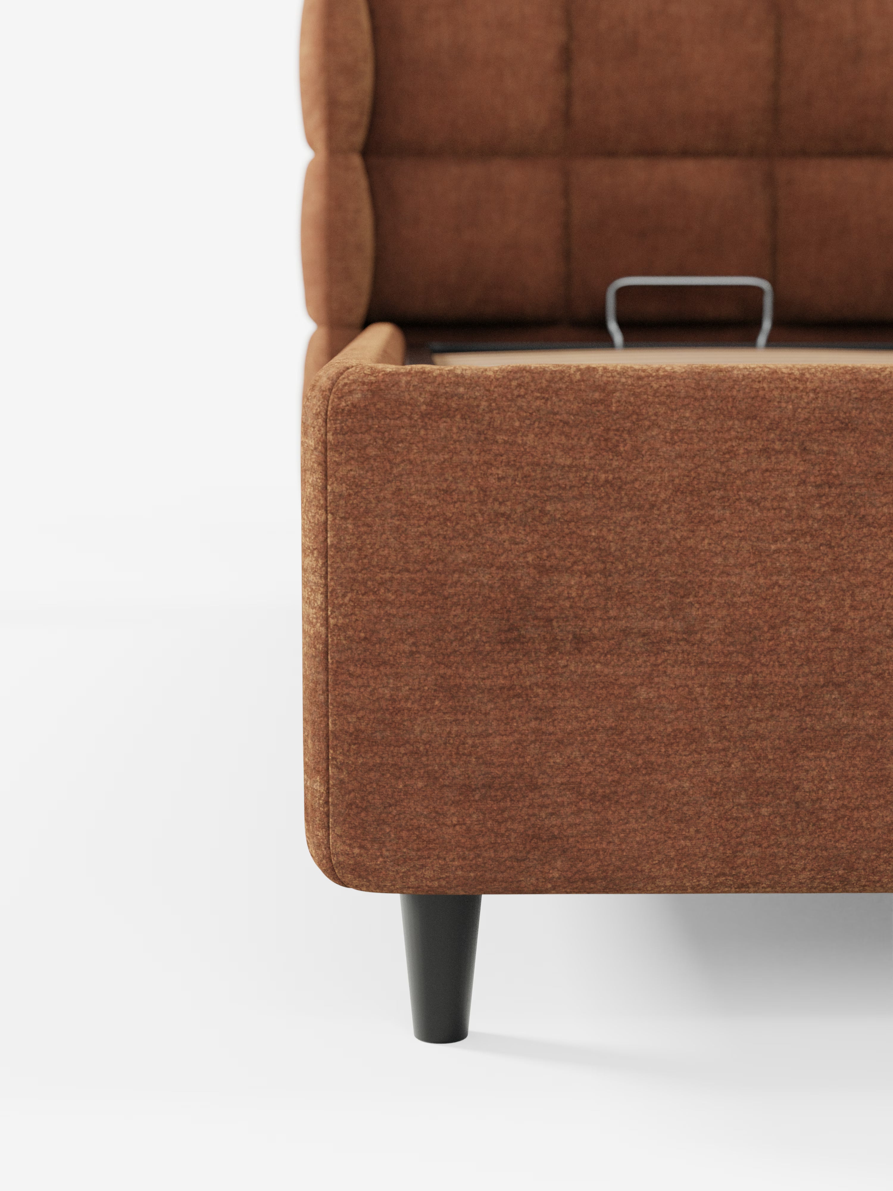 Primo piano del letto imbottito RAGNAR con contenitore e testiera capitonné avvolgente di Letti.store, rivestito in un elegante tessuto marrone certificato Oeko-Tex®. La struttura del letto presenta fianchi lisci e testurizzati e una maniglia metallica a vista nella parte superiore, poggiando su eleganti gambe angolate di colore nero. Uno sfondo bianco a tinta unita mette in risalto la struttura e i materiali di alta qualità di questo design contemporaneo.