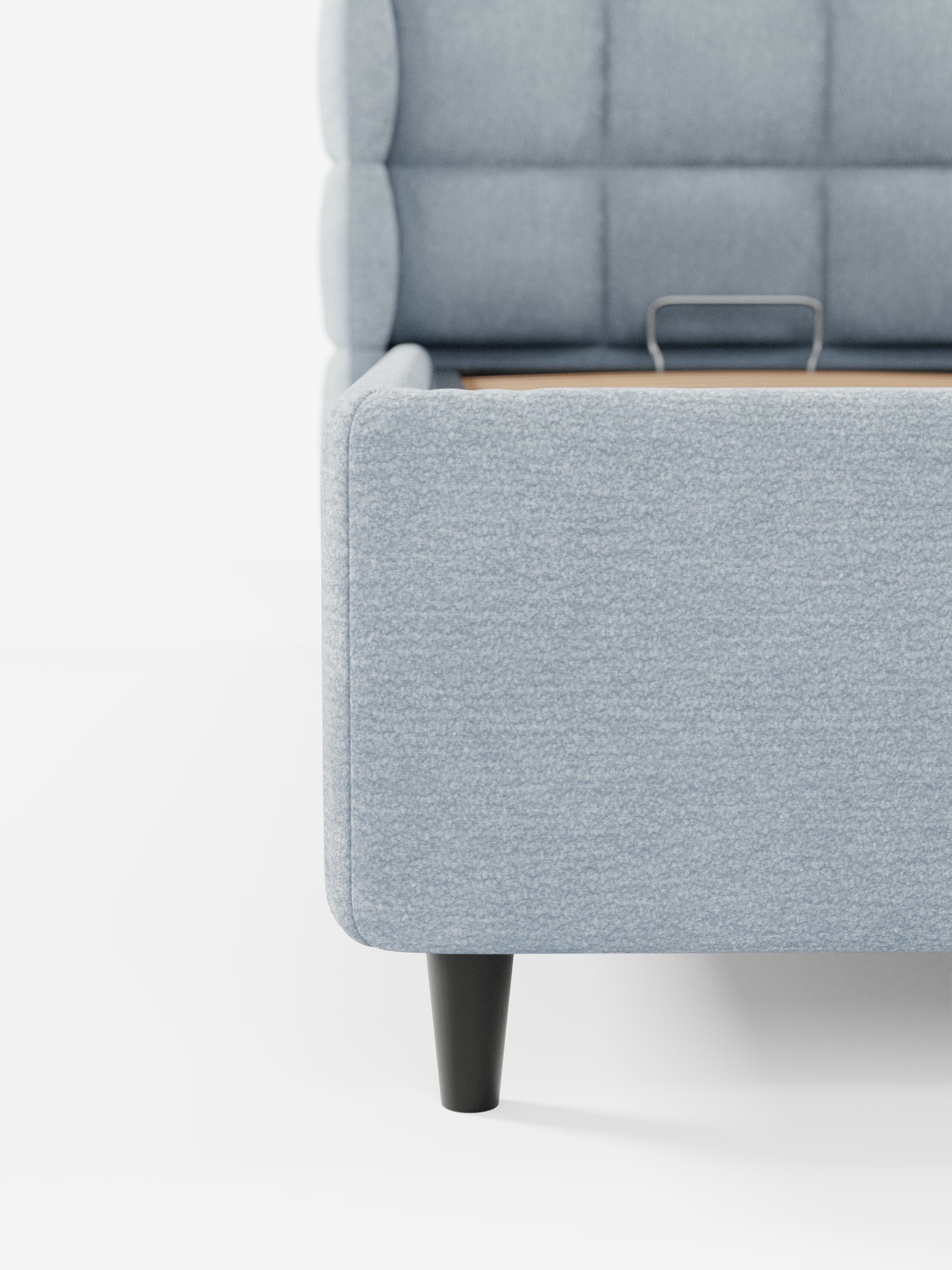 Primo piano di un moderno letto imbottito RAGNAR con contenitore e testiera capitonné avvolgente di Letti.store, caratterizzato da un elegante rivestimento in tessuto azzurro certificato Oeko-Tex® con dettagli trapuntati. Il letto poggia su gambe cilindriche nere ed è visibile una parte della base in legno con una maniglia metallica incorporata. Lo sfondo è bianco a tinta unita.