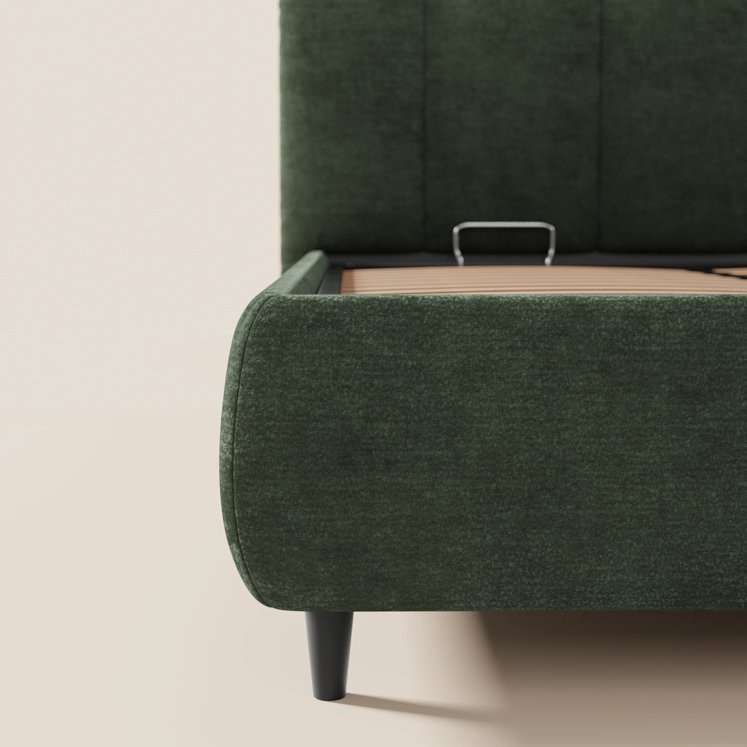 Primo piano del letto imbottito ARKAM di Letti.store, caratterizzato da una struttura in tessuto verde scuro certificato Oeko-Tex®, una testiera alta con cuciture verticali, gambe affusolate nere e una base con contenitore: perfetto per un look shabby chic in abbinamento a pareti e pavimento beige chiaro.