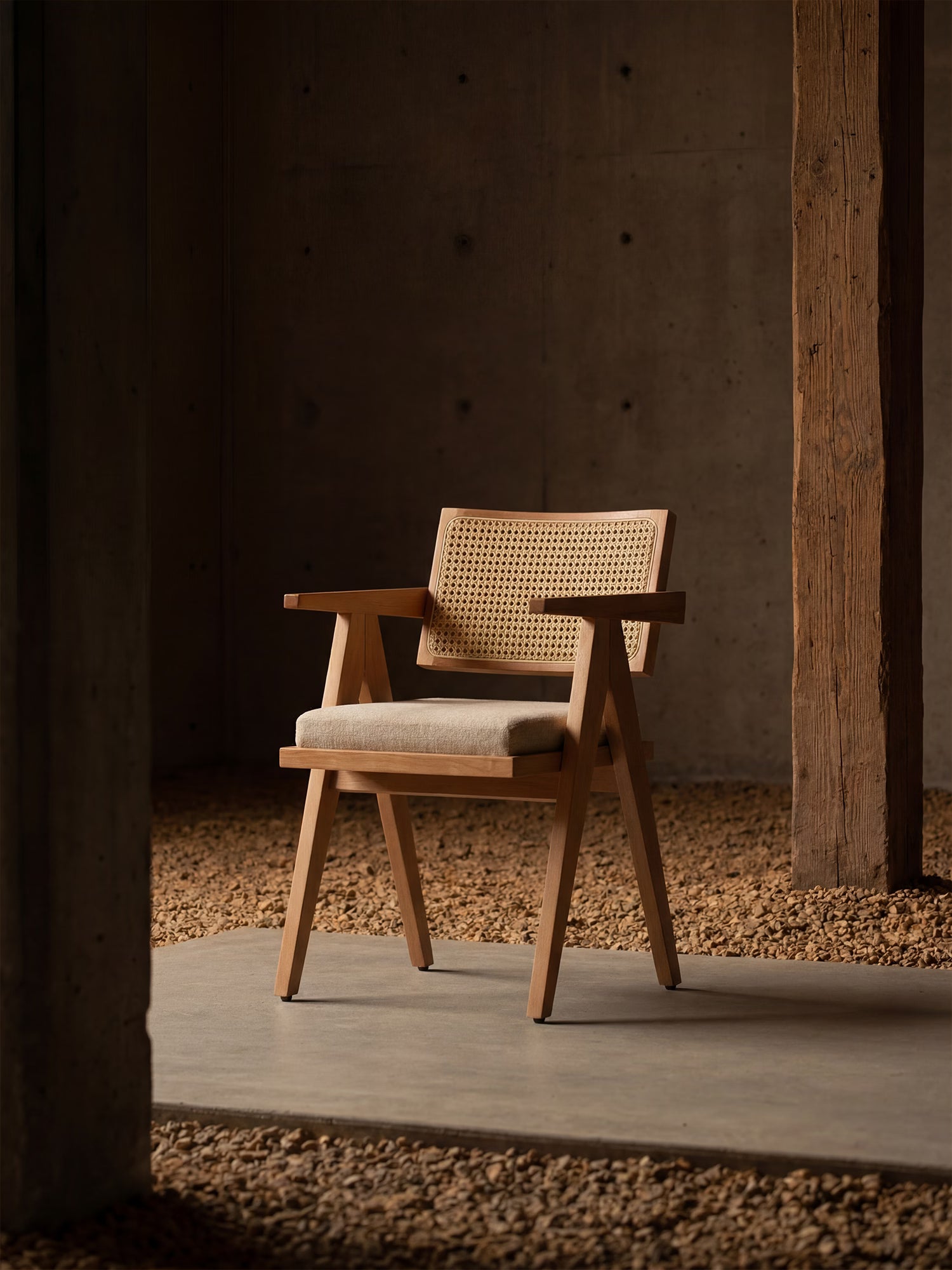 La sedia Antea, con braccioli in rovere, schienale in rattan e seduta imbottita in tessuto misto cotone di Divani.Store, è posizionata su una lastra di cemento in una stanza dallo stile minimalista e rustico, con pavimento in ghiaia, un palo di legno e una parete in cemento.