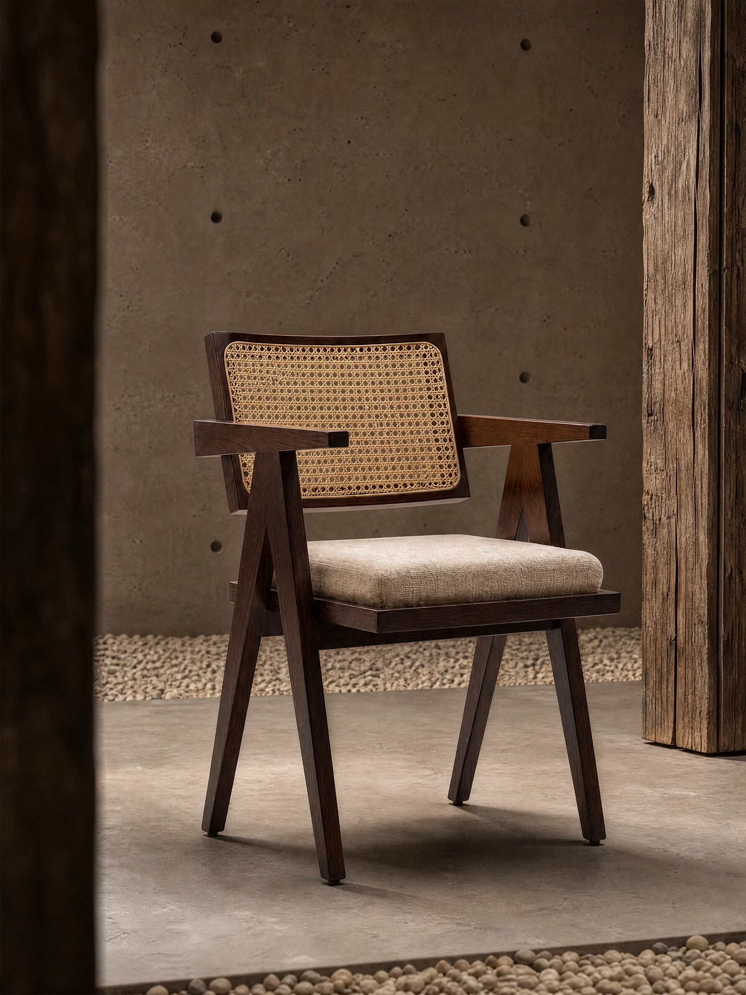 La sedia Antea con braccioli di Divani.Store, realizzata in legno massello di wengé con schienale in rattan e seduta imbottita in misto cotone, poggia su una base in cemento liscio con pareti ruvide e pilastri in legno, bordata da ciottoli bianchi.