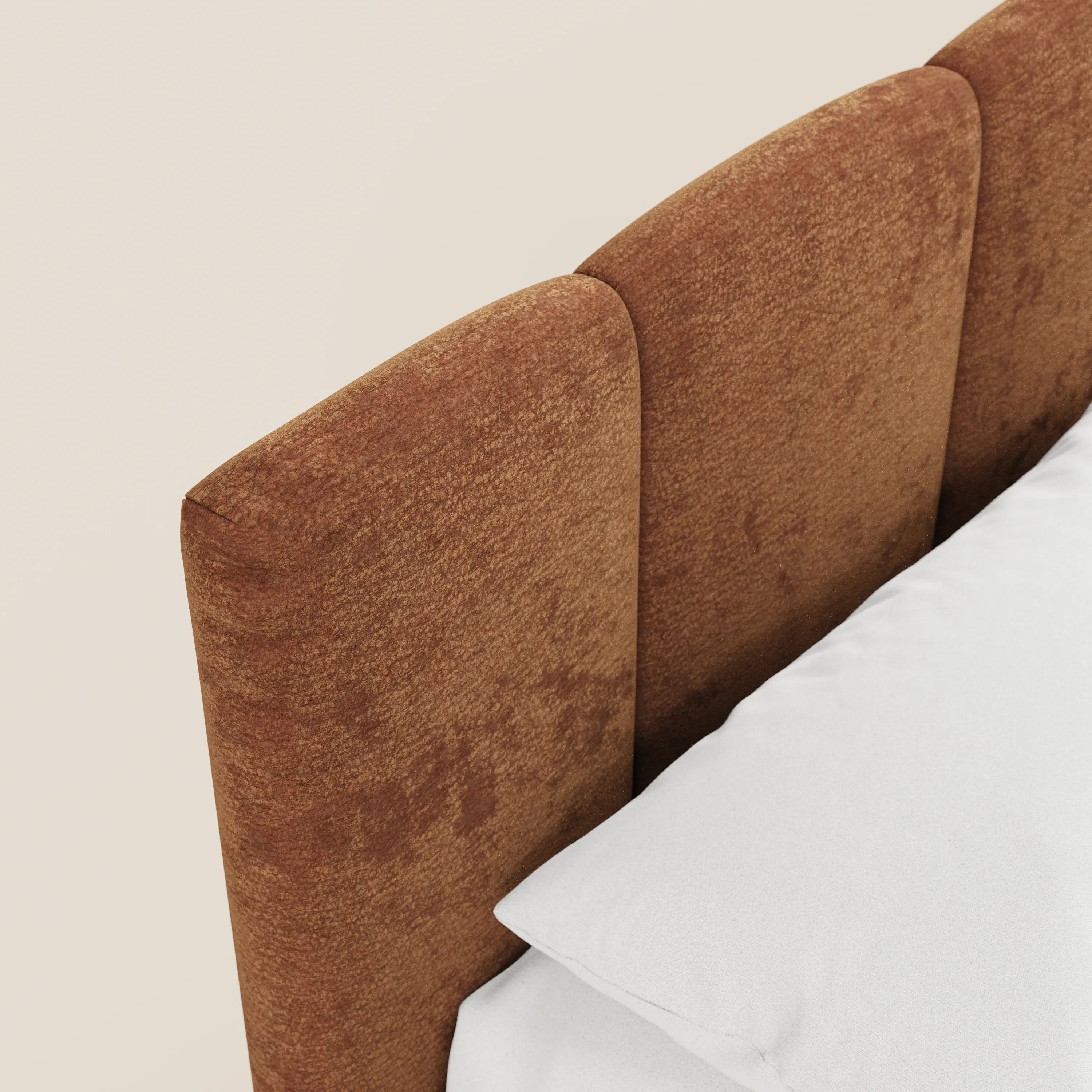 Un primo piano del letto imbottito ARKAM di Letti.store, caratterizzato da una morbida testiera color ruggine con cuciture verticali in tessuto Oeko-Tex®. Un cuscino bianco e un lenzuolo fanno da contrappunto alla parete beige, conferendo all’insieme un tocco shabby chic.