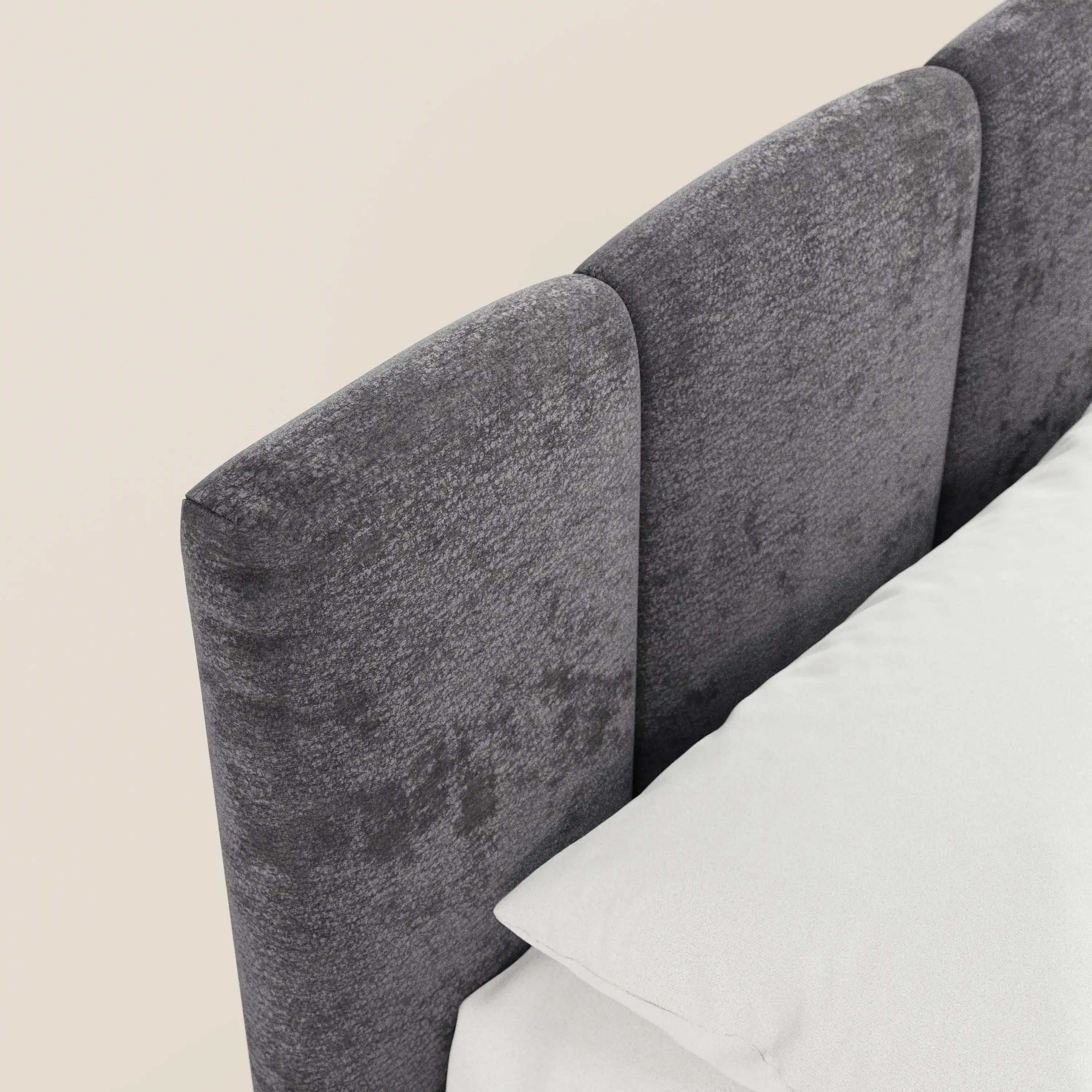 Un primo piano di ARKAM di Letti.store, un letto imbottito in stile shabby chic con contenitore e testiera verticale in tessuto Oeko-Tex®. La testiera grigio scuro presenta tre pannelli arrotondati, con biancheria da letto e cuscini bianchi su uno sfondo beige.