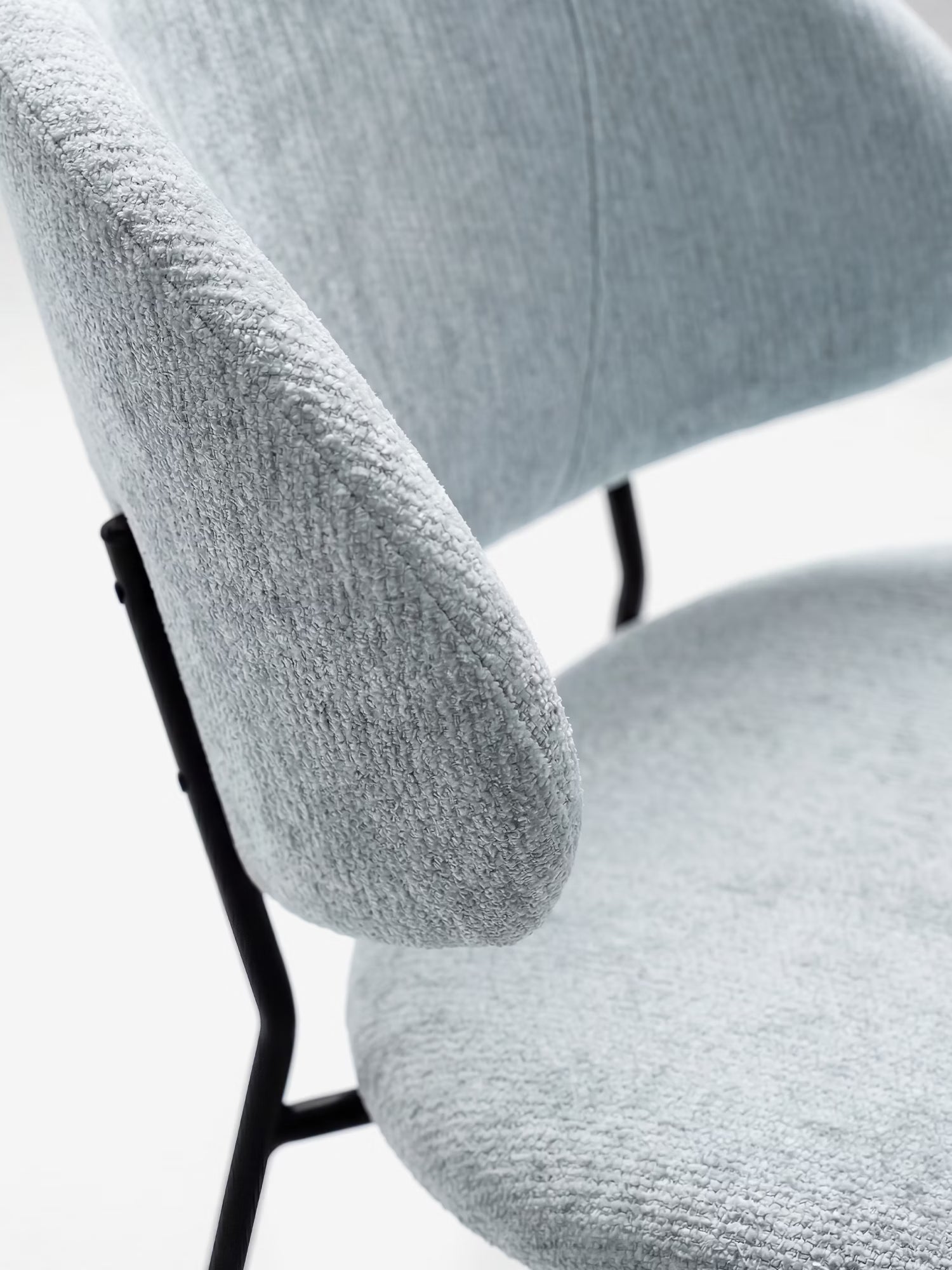 Primo piano della sedia Bruma di Divani.Store, caratterizzata da un elegante tessuto goffrato in grigio chiaro, un design minimalista con schienale avvolgente, imbottitura morbida e robuste gambe in metallo nero. Sfondo bianco a tinta unita.