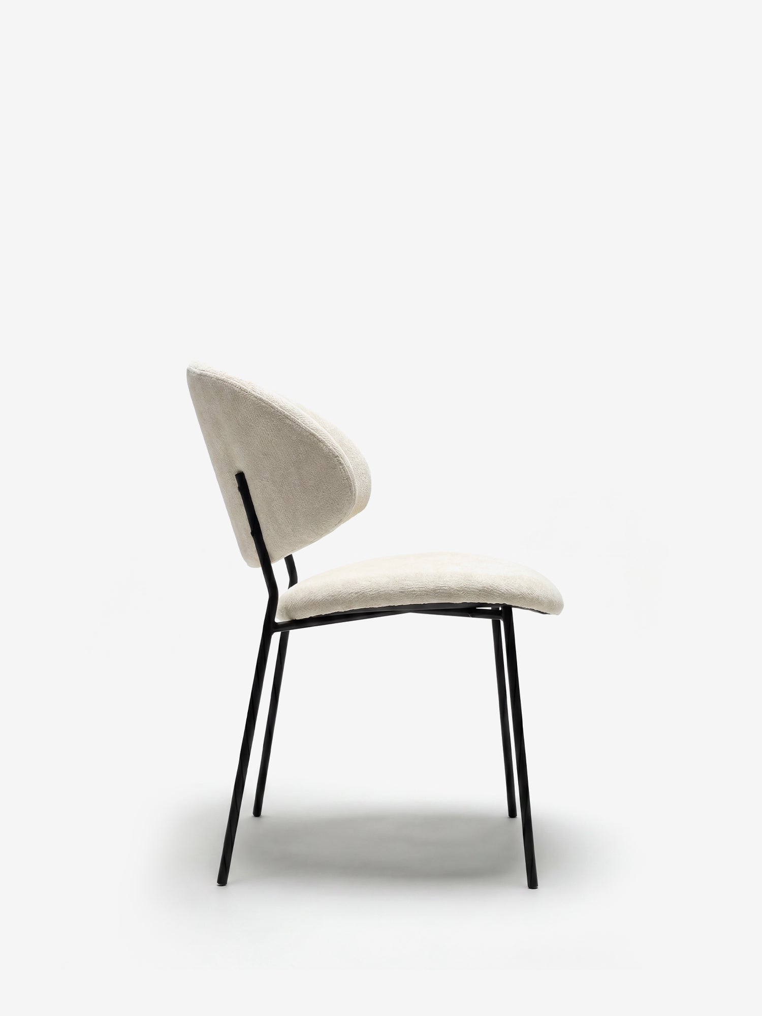 Vista laterale della sedia Bruma di Divani.Store, caratterizzata da un elegante tessuto goffrato color beige chiaro e da uno schienale avvolgente, con quattro gambe sottili in metallo nero inclinate verso l’esterno: un design minimalista messo in risalto da uno sfondo bianco a tinta unita.