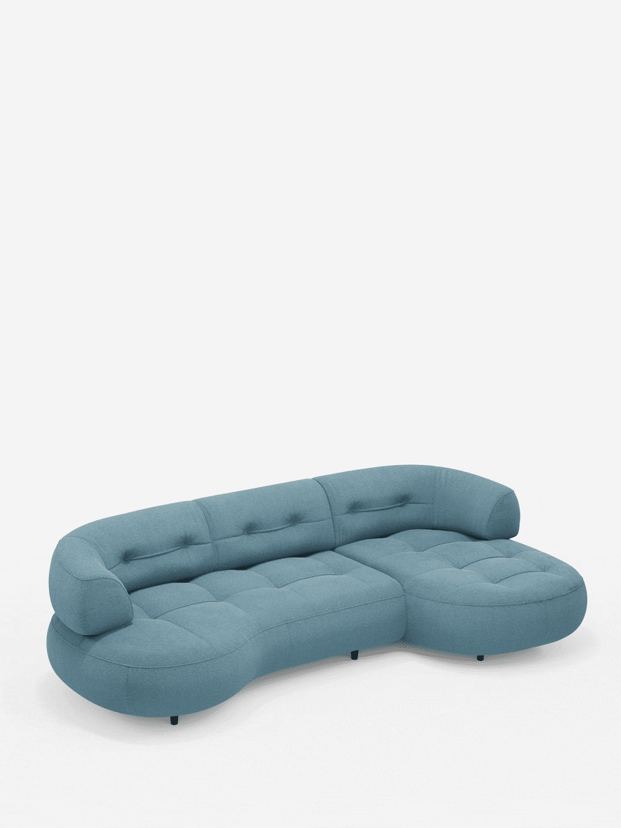 Un divano angolare Croissant dalla forma ricurva a fagiolo, con schienali scorrevoli in morbido tessuto effetto cotone smacchiabile T33, rivestito in tessuto blu polveroso con seduta e schienale trapuntati e piedini bassi scuri su sfondo bianco.