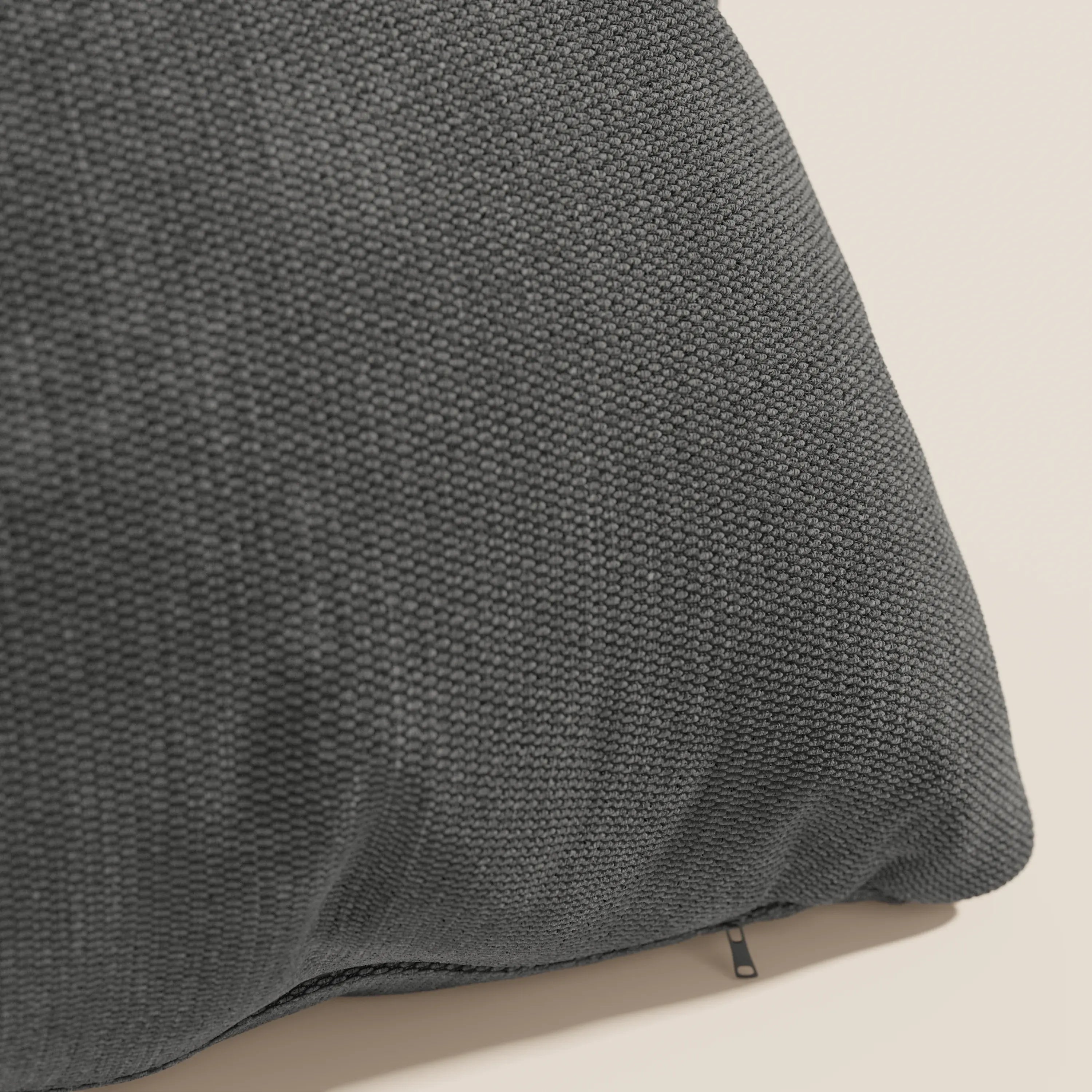 Un primo piano del cuscino Miami di Divani.Store in T14 antracite mostra il suo tessuto grigio scuro, strutturato e resistente all'acqua, e la sua discreta cerniera; lo sfondo è un semplice beige chiaro.