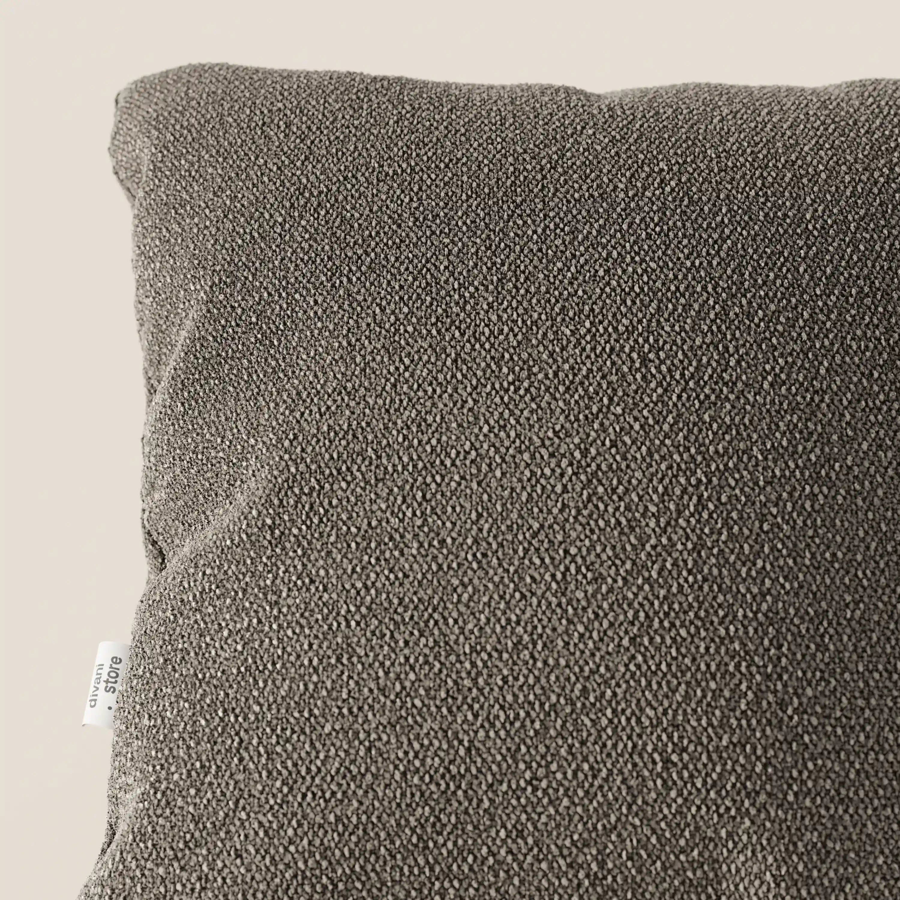 Primo piano dell'angolo superiore sinistro di un cuscino Lambada grigio scuro di Divani.Store, realizzato in tessuto simil-cotone fresco, antimacchia e impermeabile (T33), con una piccola etichetta bianca sul bordo, su uno sfondo beige chiaro.