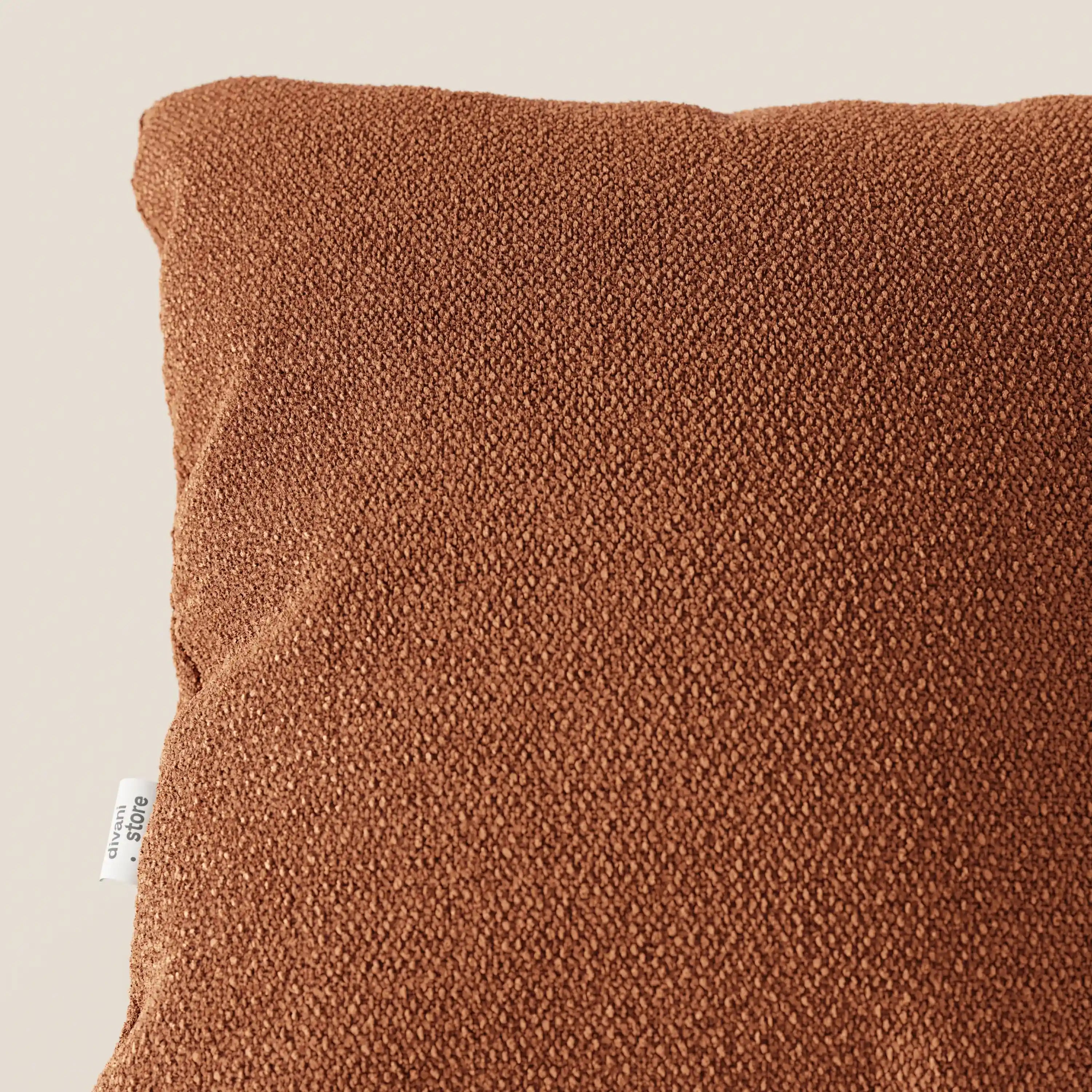 Primo piano dell'angolo superiore sinistro di un cuscino Lambada marrone di Divani.Store, realizzato in tessuto smacchiabile e impermeabile tipo cotone naturale. Una piccola etichetta bianca con testo nero è visibile sul bordo sinistro del cuscino su uno sfondo neutro.