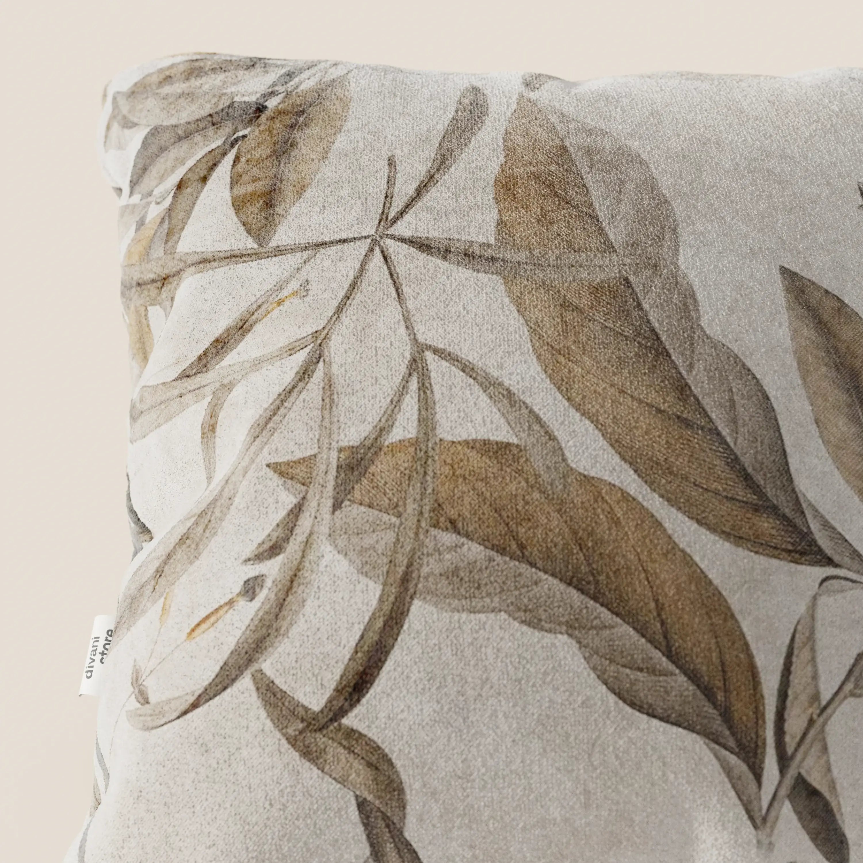 Primo piano del cuscino Lemon elegante a fantasia in velluto impermeabile beige di Divani.Store, caratterizzato da grandi foglie botaniche marroni e abbronzate su sfondo beige chiaro, texture vellutata e una piccola etichetta bianca sulla cucitura sinistra.
