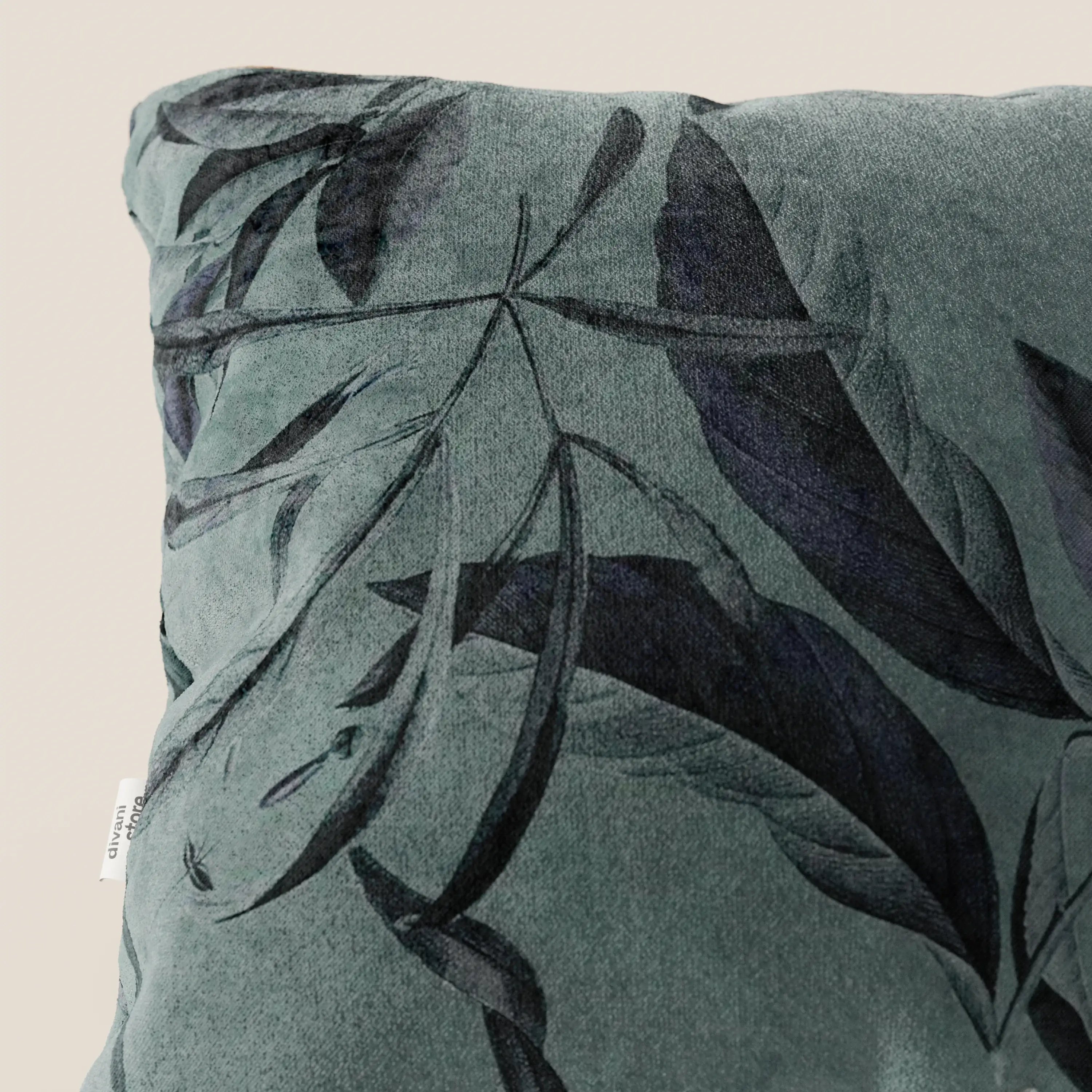 Un primo piano della collezione Lemon elegante di cuscini a fantasia in velluto impermeabile petrolio di Divani.Store mostra un morbido cuscino verde con un sottile motivo a foglie scure e un'etichetta bianca visibile, su uno sfondo beige.