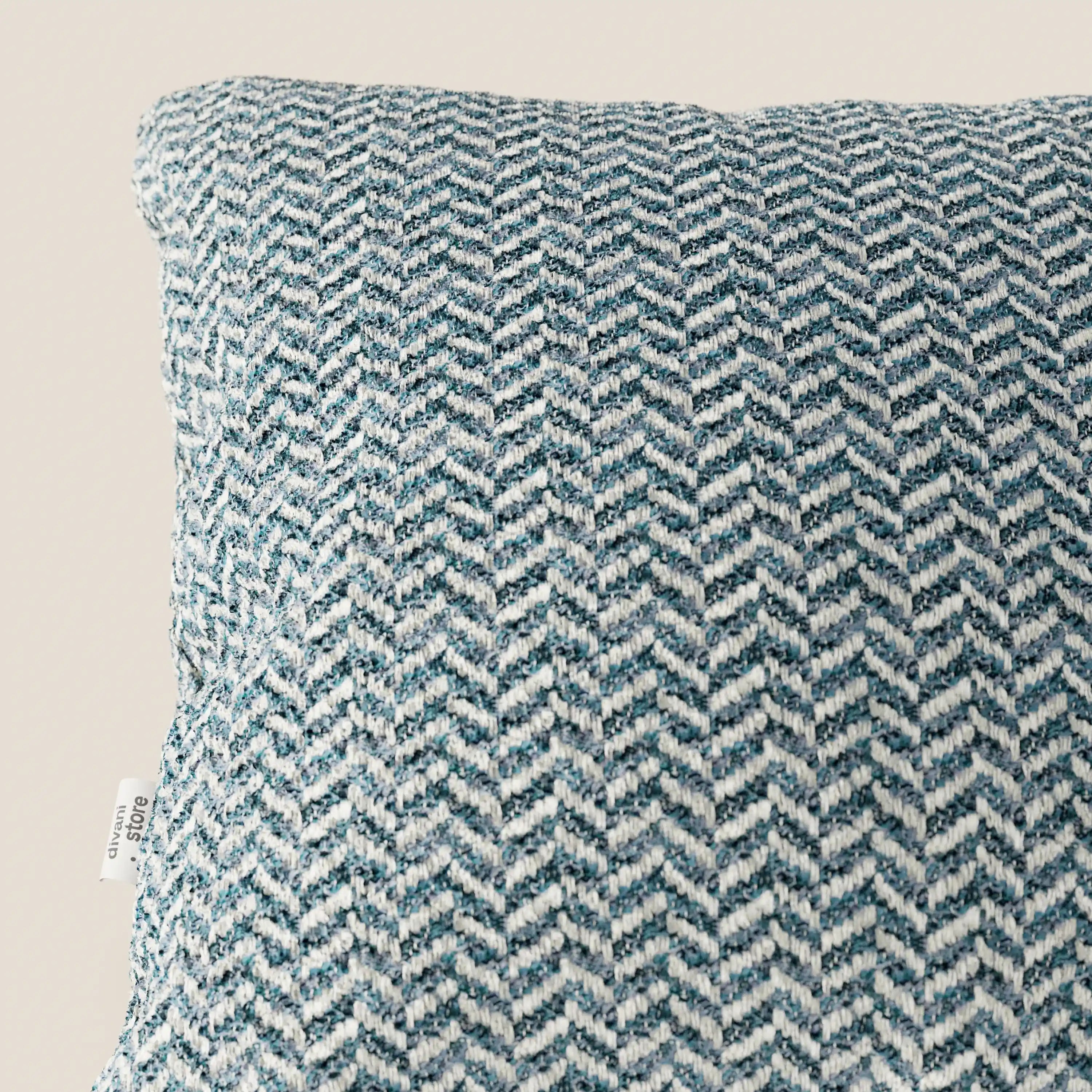 Primo piano del cuscino Sorrento 45x45 in lino di Divani.Store, realizzato in tessuto testurizzato nei toni del verde acqua e del bianco con motivo a chevron intrecciato e una piccola etichetta bianca sul bordo sinistro, su uno sfondo chiaro a tinta unita. Cuscino lavabile in lavatrice.