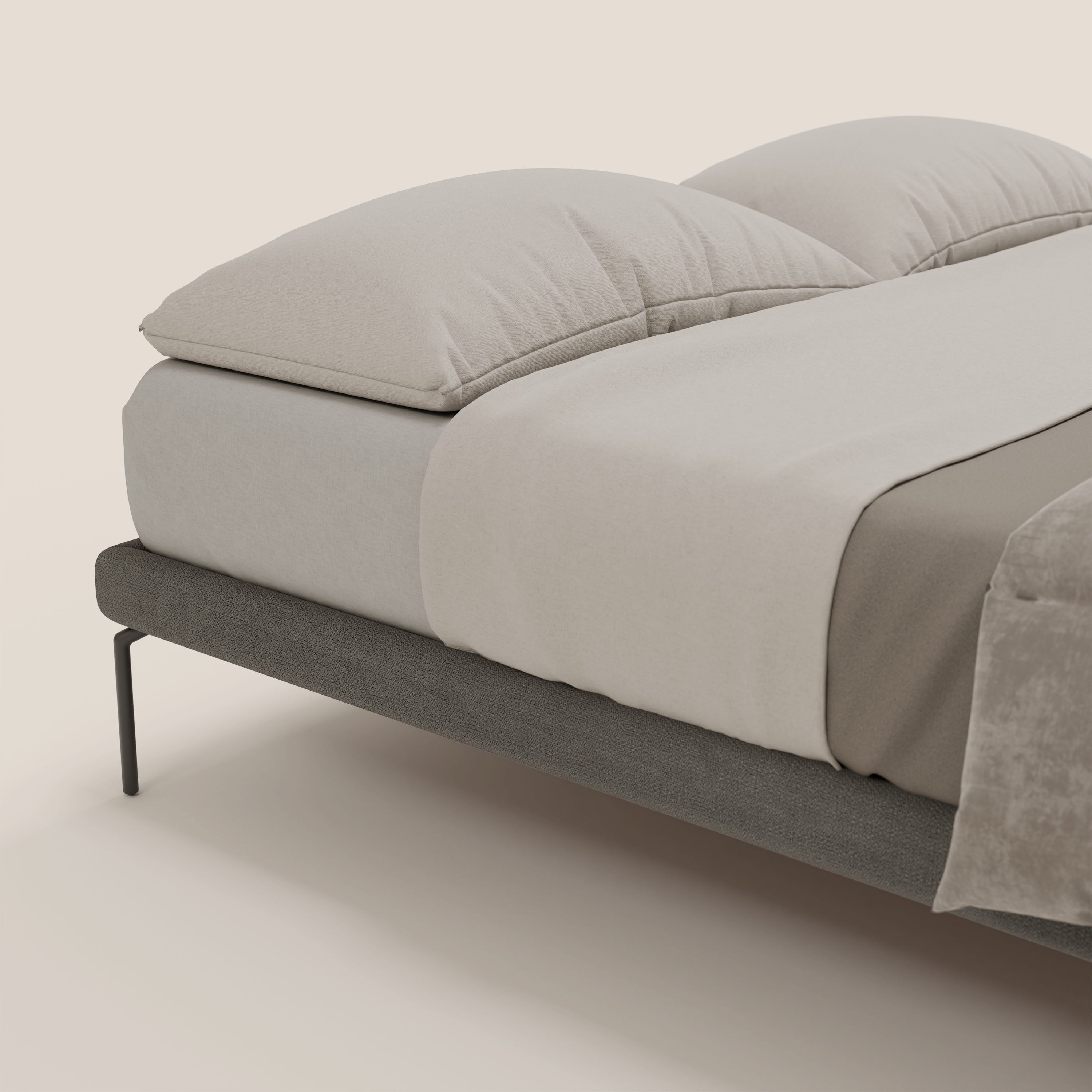 Il sommier sfoderabile Ring di Letti.store è caratterizzato da gambe nere alte 24 cm e da una struttura in tessuto impermeabile T14 grigio. La biancheria da letto beige chiaro con due grandi cuscini e una coperta ripiegata color tortora completano il look su un morbido sfondo crema.