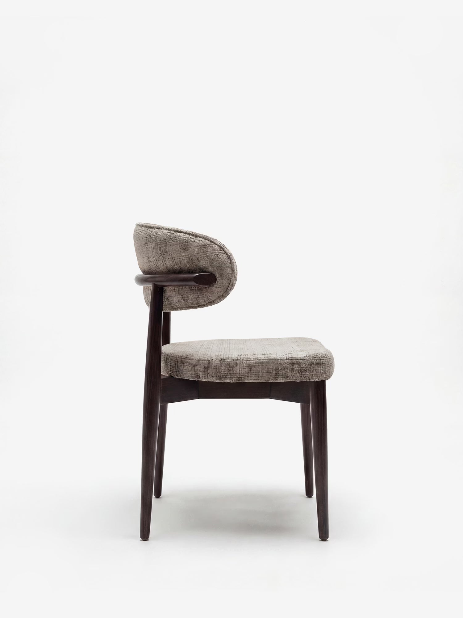 La sedia di design imbottita Dafne di Divani.Store presenta una struttura in legno massello di wengé, uno schienale imbottito e curvo e una seduta in tessuto ciniglia OEKO-TEX® di colore grigio neutro, qui raffigurata di profilo su uno sfondo bianco a tinta unita.