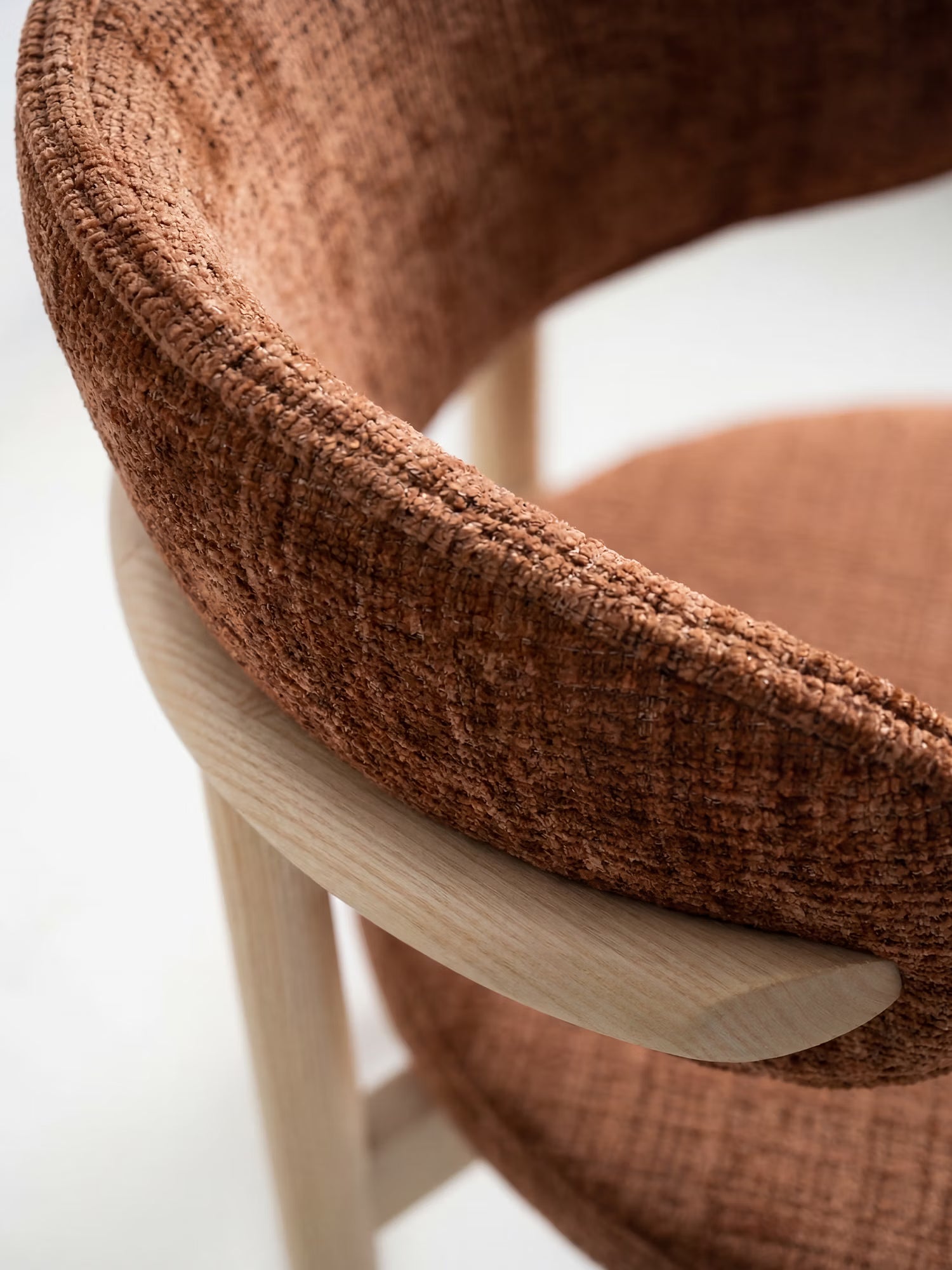 Un primo piano della sedia imbottita Dafne di Divani.Store mette in risalto il suo rivestimento in ciniglia OEKO-TEX® dai toni ruggine e l'elegante schienale curvo, abbinati a braccioli e gambe in rovere massello naturale su uno sfondo bianco.