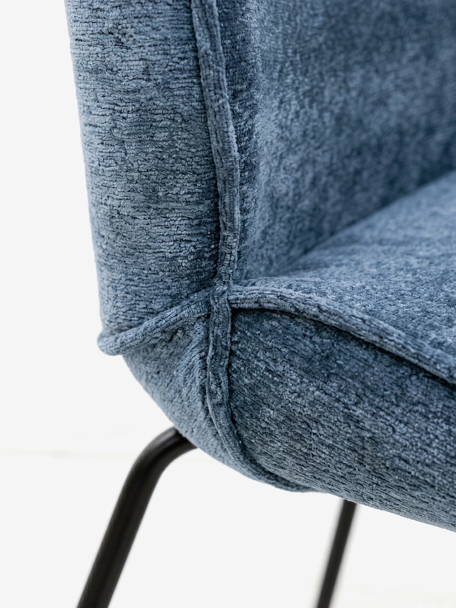 Primo piano della sedia imbottita Iride di Divani.Store, caratterizzata da un tessuto materico blu morbido al tatto, cuciture a vista sui bordi e una struttura con gambe in metallo nero. L'attenzione è focalizzata sul punto di incontro tra seduta e schienale, su uno sfondo chiaro e sfocato.