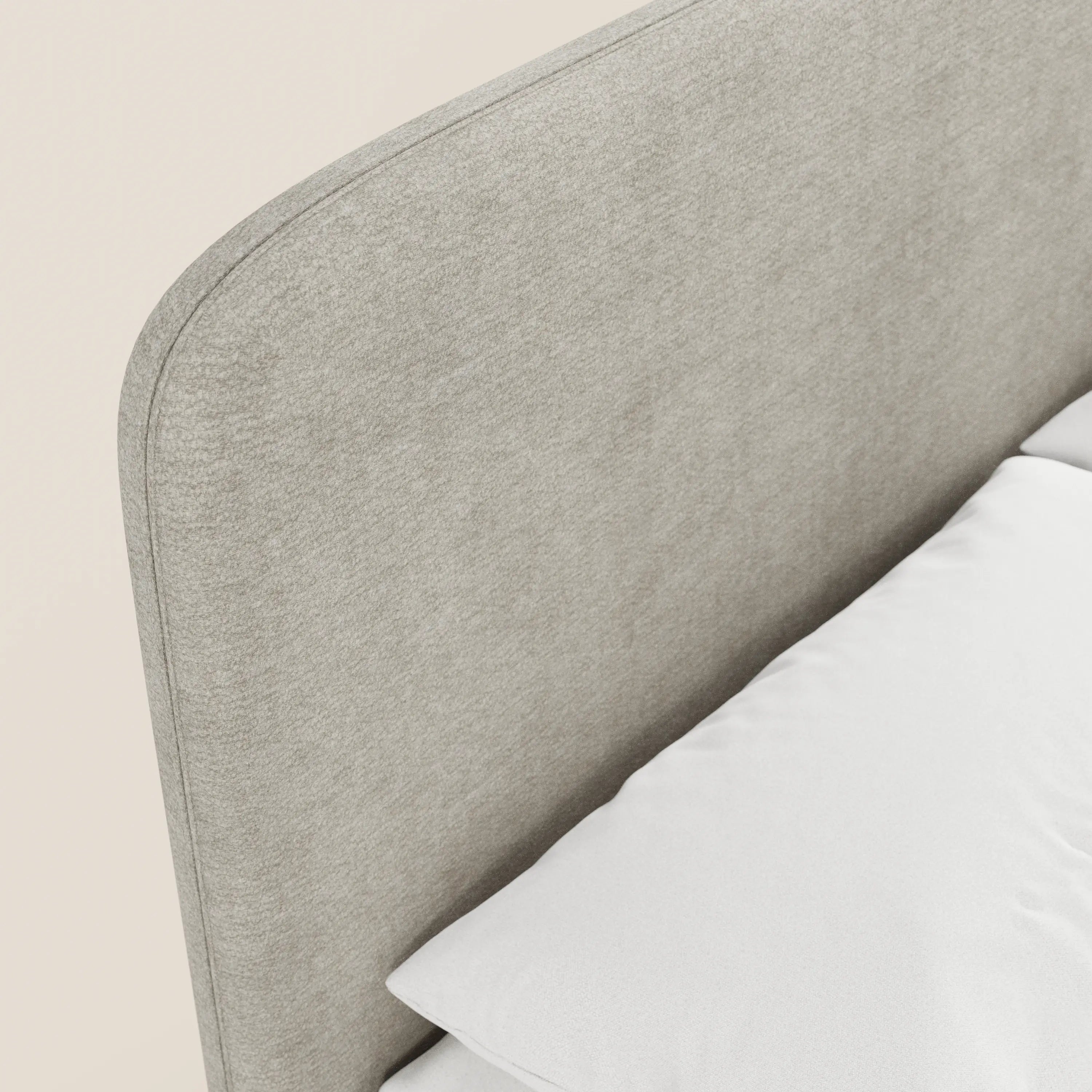 Primo piano del letto imbottito KAELOS Letto Minimal di Letti.store, caratterizzato da una testiera in tessuto ciniglia grigio chiaro con angoli arrotondati, gambe alte e vano contenitore. Presentato con biancheria da letto bianca su uno sfondo color crema per esaltarne lo stile morbido e minimalista.
