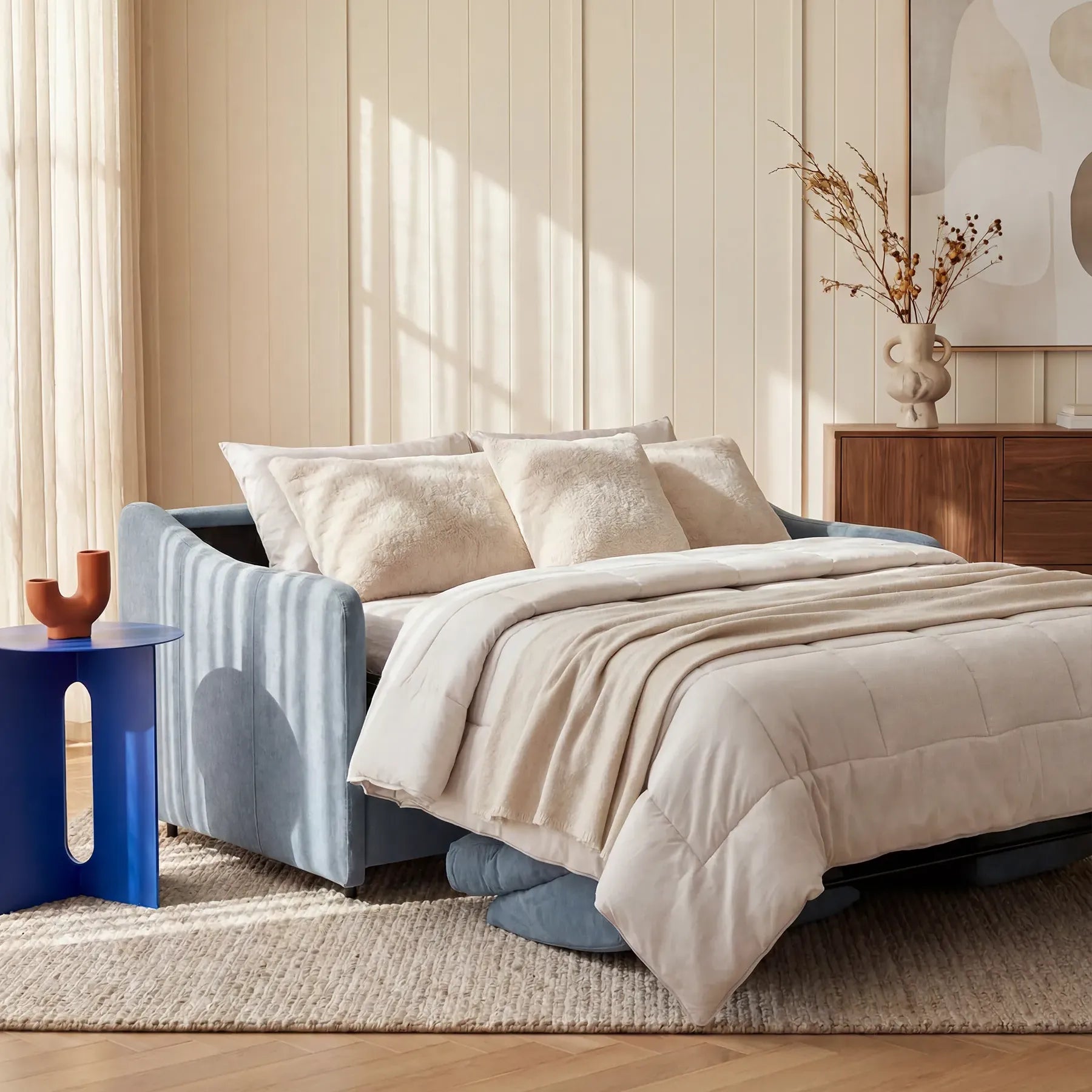 Un soggiorno moderno è arredato con il divano letto blu a 3 posti “Lodovico” di Divani.Store, aperto con biancheria da letto e cuscini beige. Accanto si trova un tavolino blu con un vaso marrone; la luce del sole filtra attraverso le tende trasparenti, illuminando una cassettiera in legno e un quadro astratto.