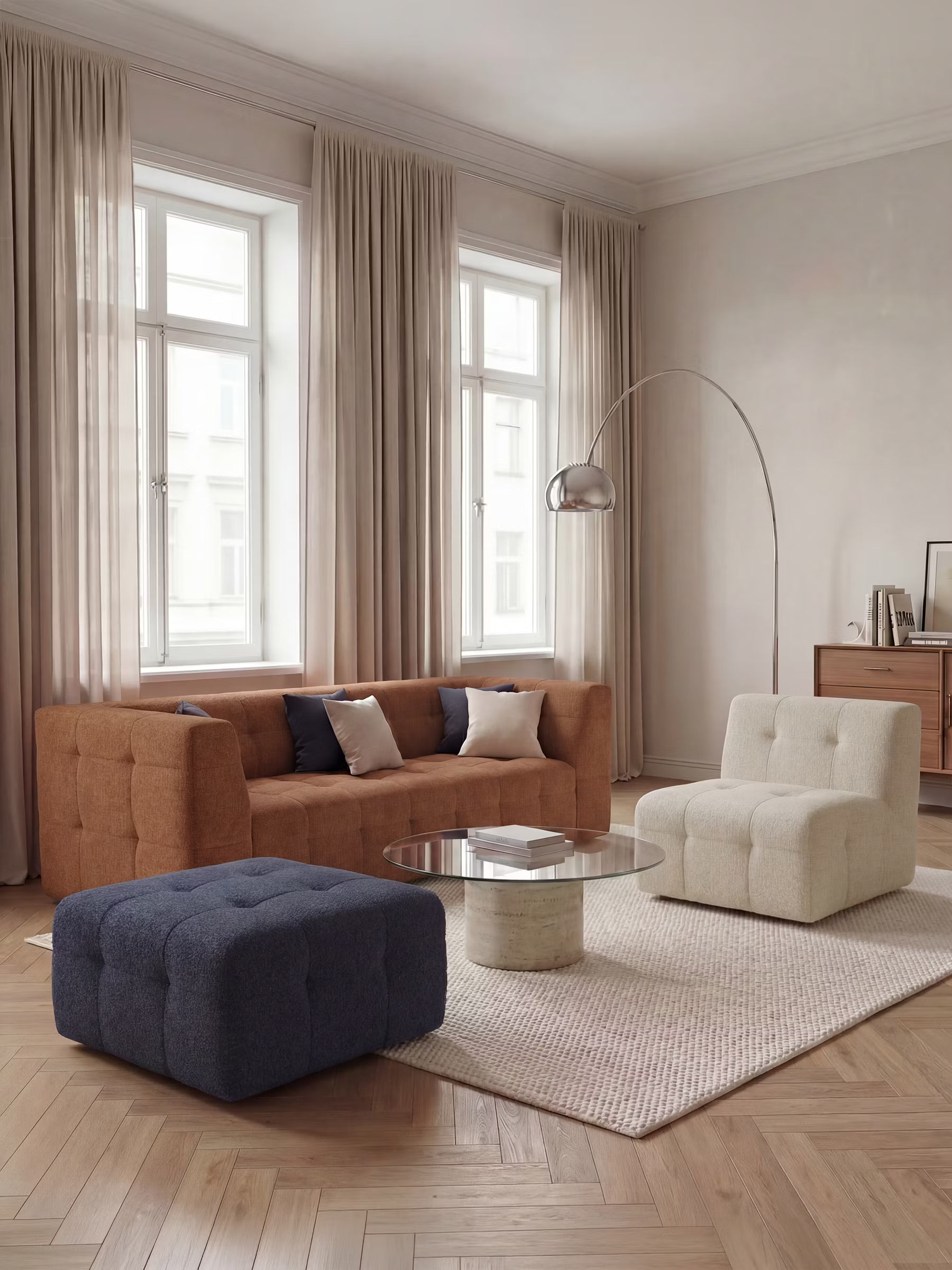 Soggiorno moderno con divano modulare Mosaic Divano a 3 e 4 posti in bouclé: imbottito e dal design in tessuto antimacchia T21 di Divani.Arredamento nei toni del marrone, poltrona color crema, pouf blu navy, tavolino in vetro, tappeto beige, pavimento in parquet, lampada da terra ad arco, credenza, libri e ampie finestre con tende trasparenti. La palette di colori è neutra e calda.