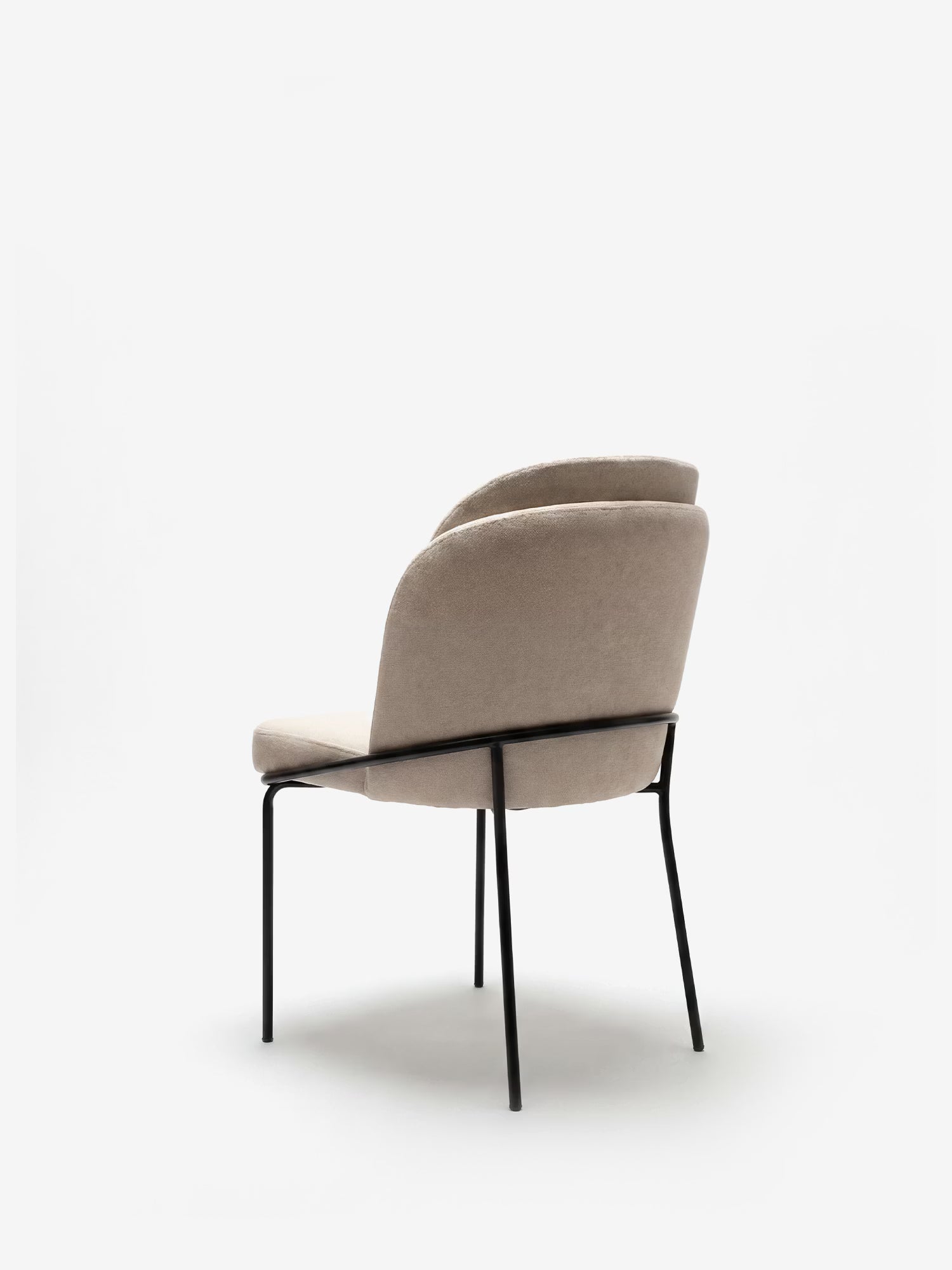 La sedia imbottita Plissè in tessuto impermeabile di Divani.Store presenta uno schienale beige plissettato e sottili gambe in metallo nero; è raffigurata in un’inquadratura a tre quarti posteriore su uno sfondo bianco a tinta unita.