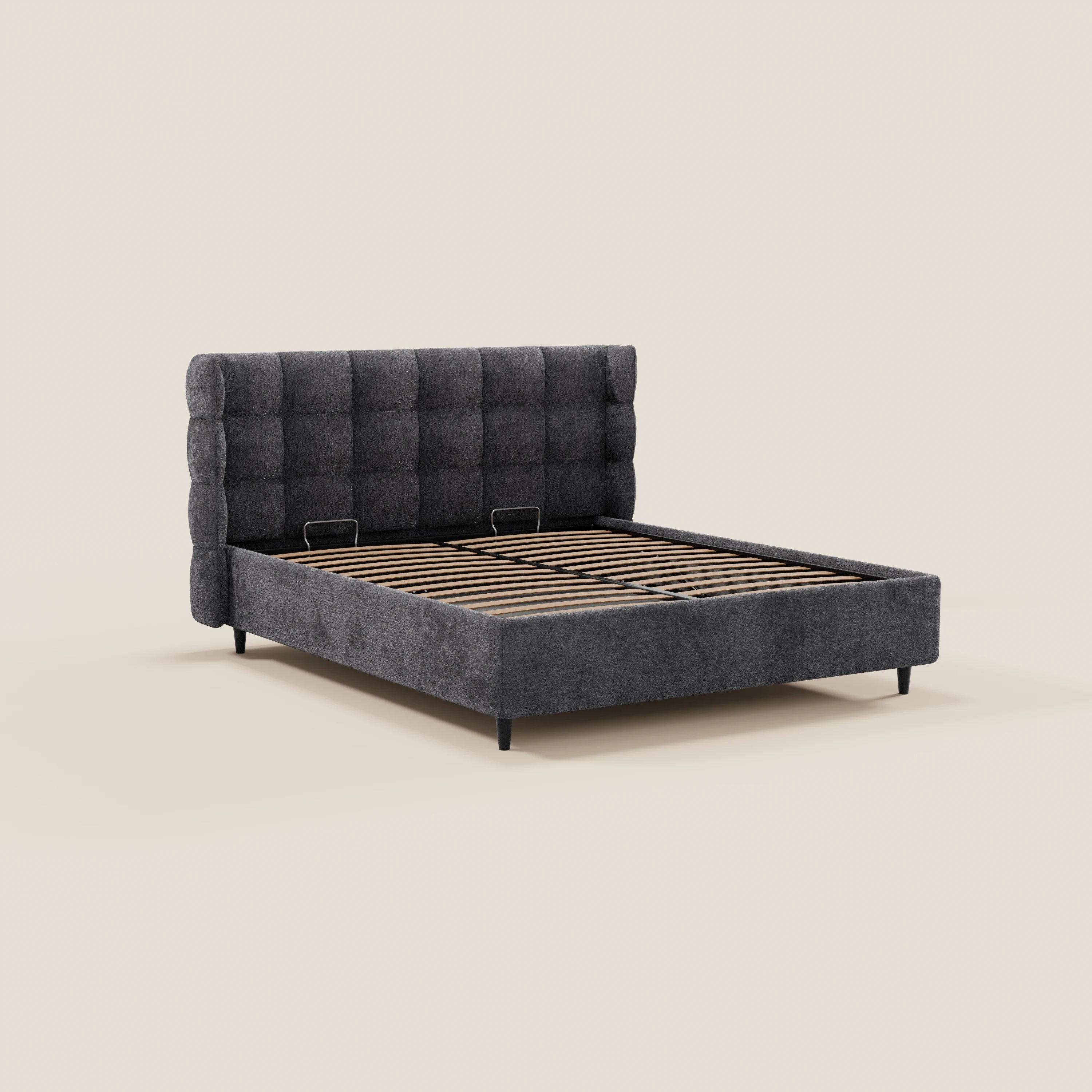 Il letto imbottito RAGNAR di Letti.store presenta una testiera alta e avvolgente con rivestimento capitonné in tessuto grigio scuro certificato Oeko-Tex®, gambe di sostegno in metallo e doghe in legno. Il letto è vuoto, senza materasso né biancheria da letto, e viene mostrato su uno sfondo beige a tinta unita.